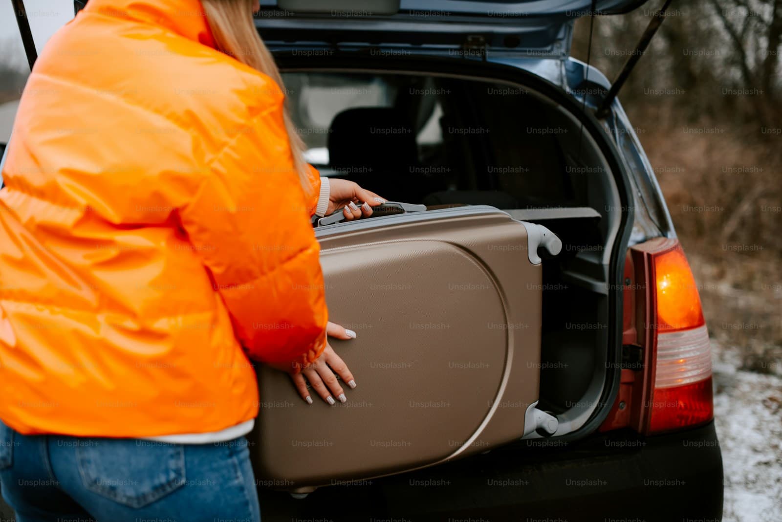 Person in an orange jacket loading a suitcase into a car trunk on snowy ground