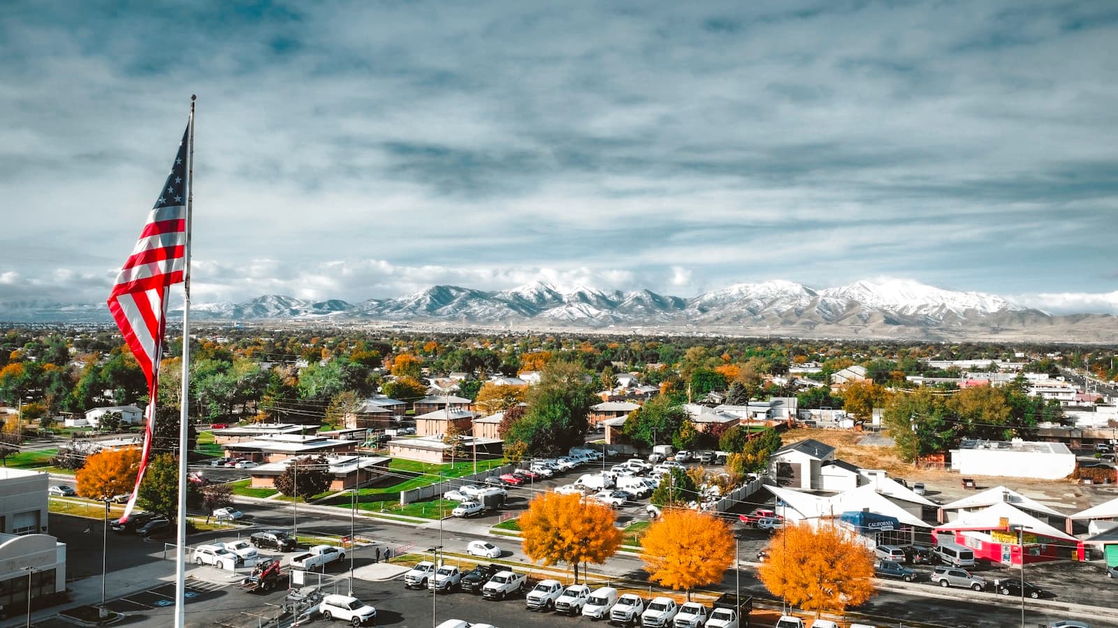 American flag flying over small town with snow-capped mountains in background and autumn trees below