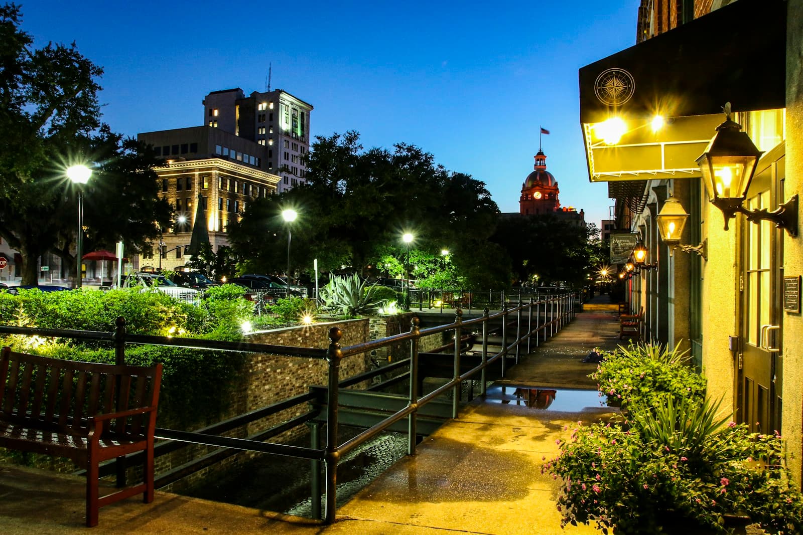 Illuminated evening walkway with railings and gardens, historic buildings and church dome in background