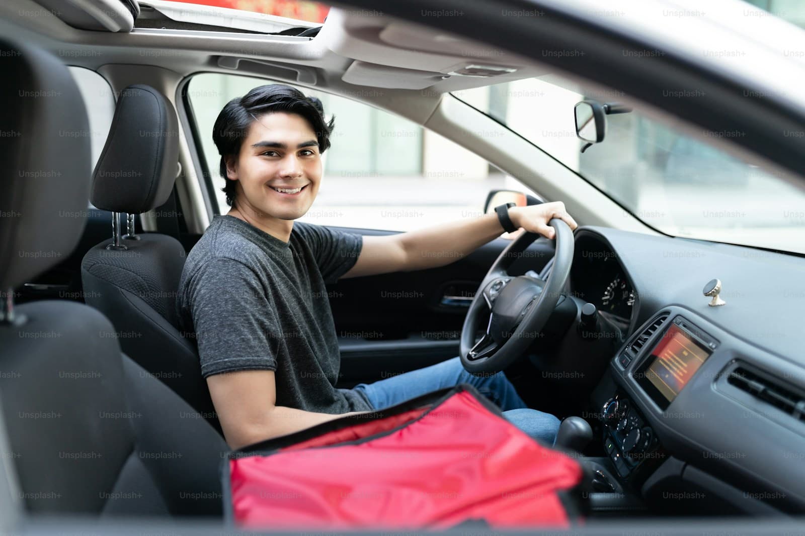 Young driver smiling at the wheel with a red delivery bag on the passenger seat
