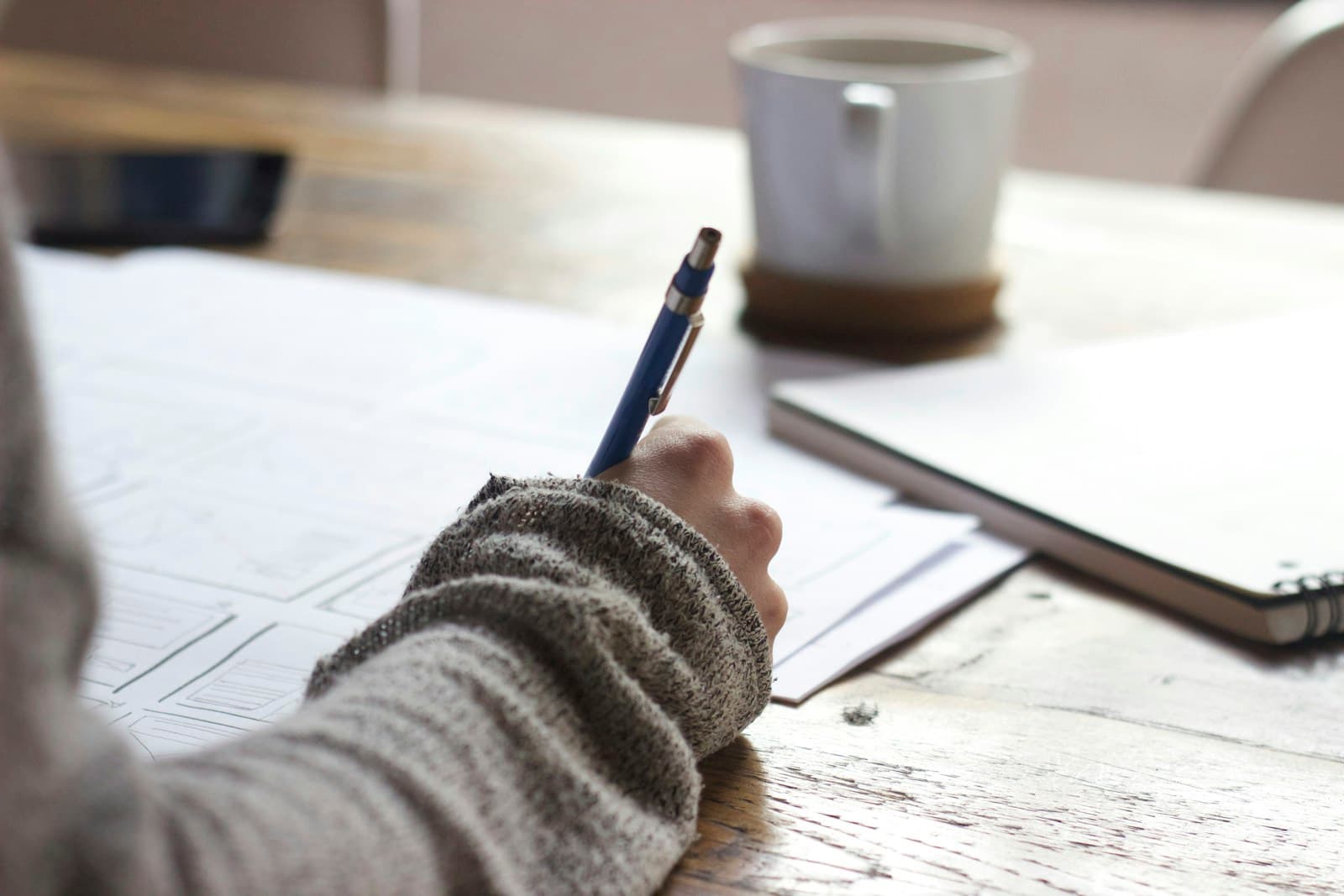 Person in a knitted sweater writing on papers beside a coffee mug and tablet