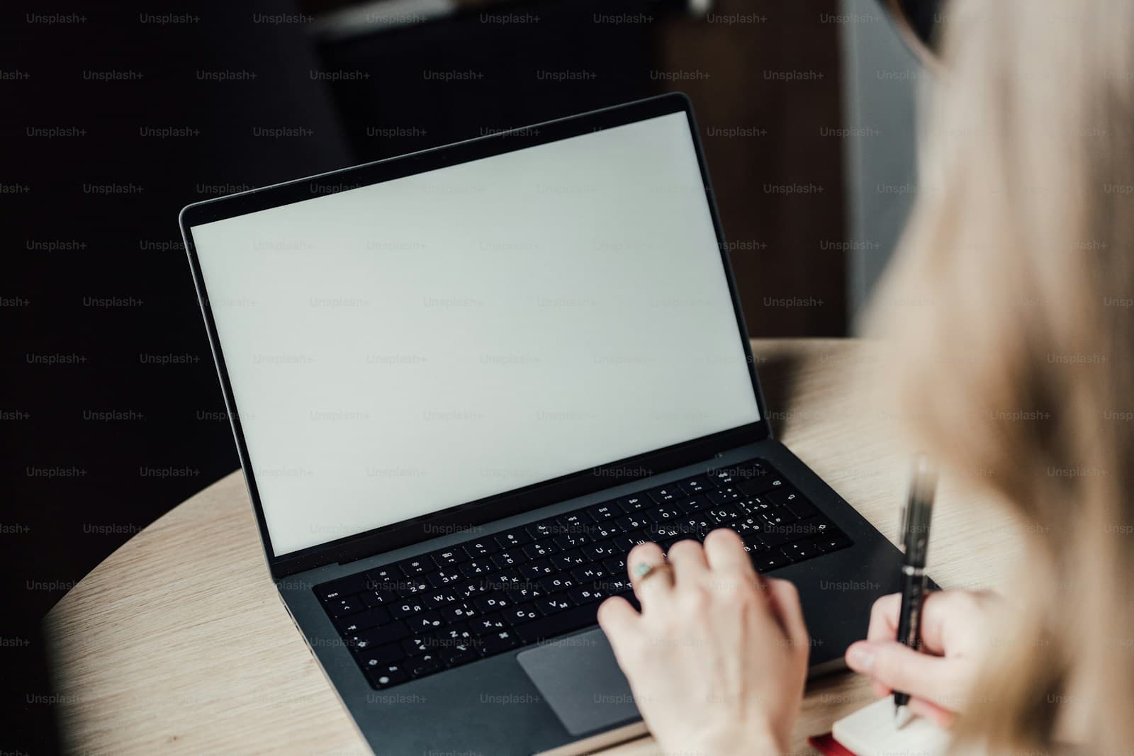 Person with blonde hair typing on a black laptop while holding a pen