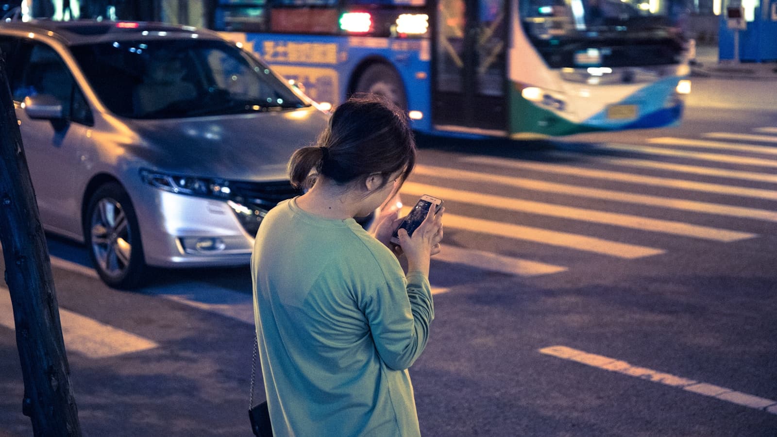 Woman in a green sweater on her phone on a city street at night as traffic passes