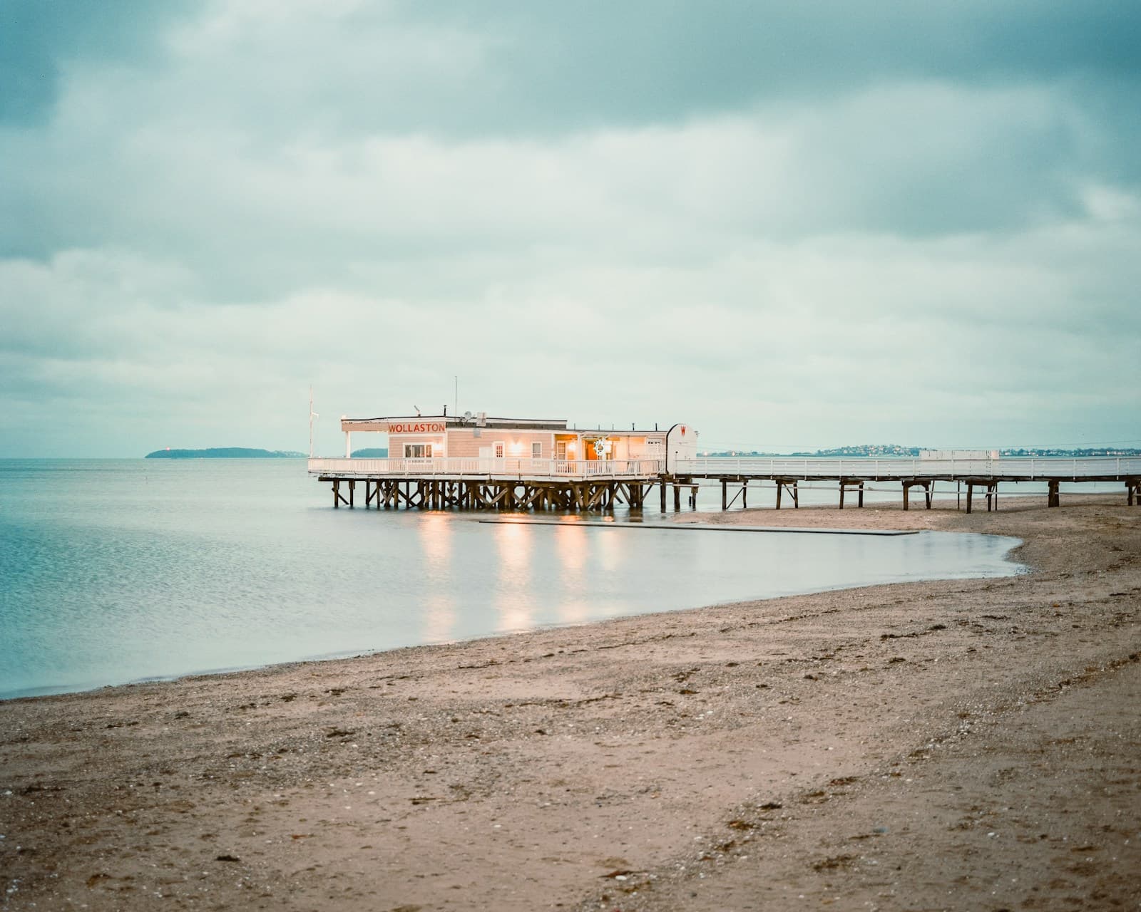 Wooden pier extending into calm water with small building, sandy beach and cloudy sky