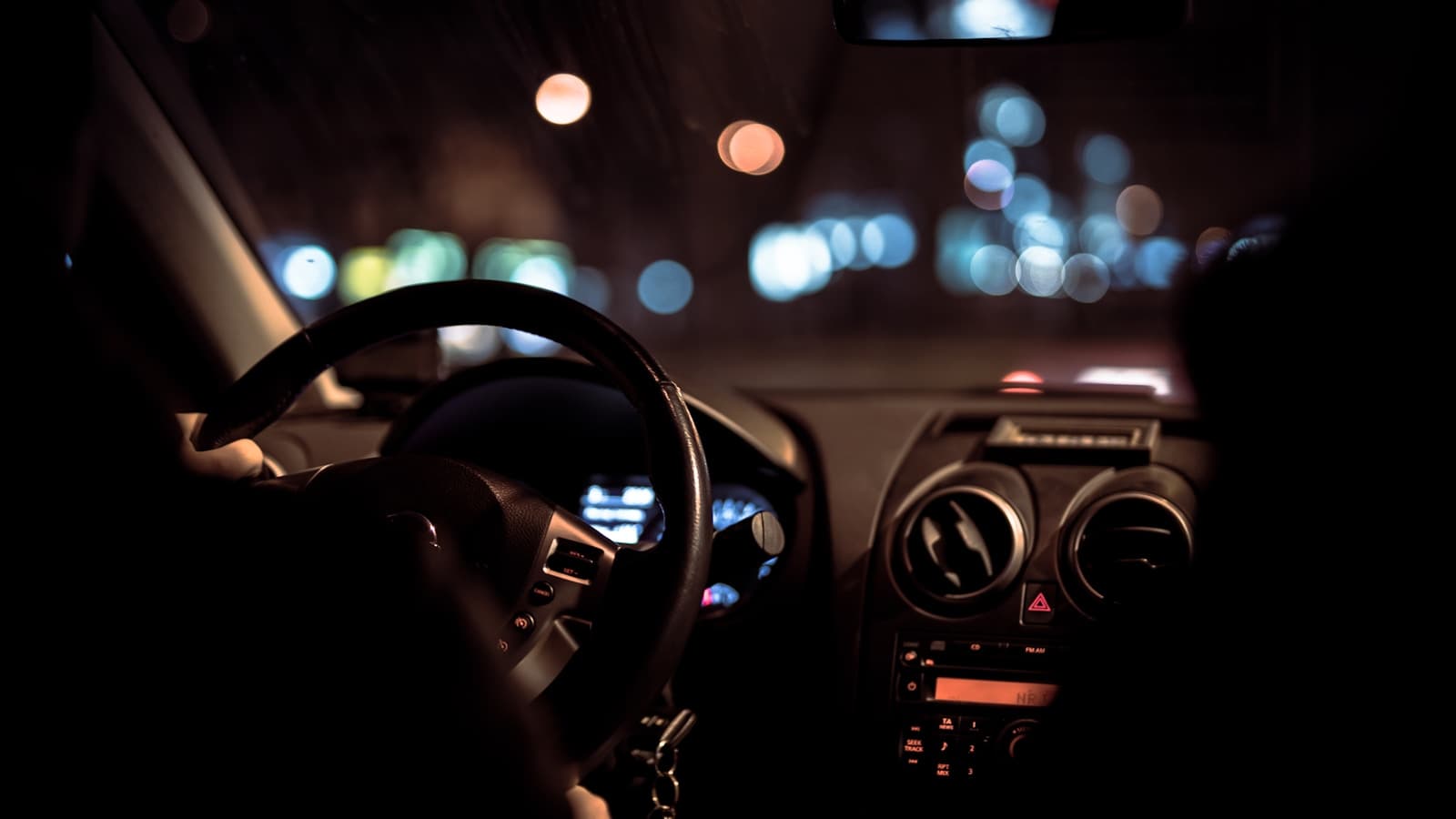 Driver's hands on steering wheel at night with city lights visible through windshield and illuminated dashboard