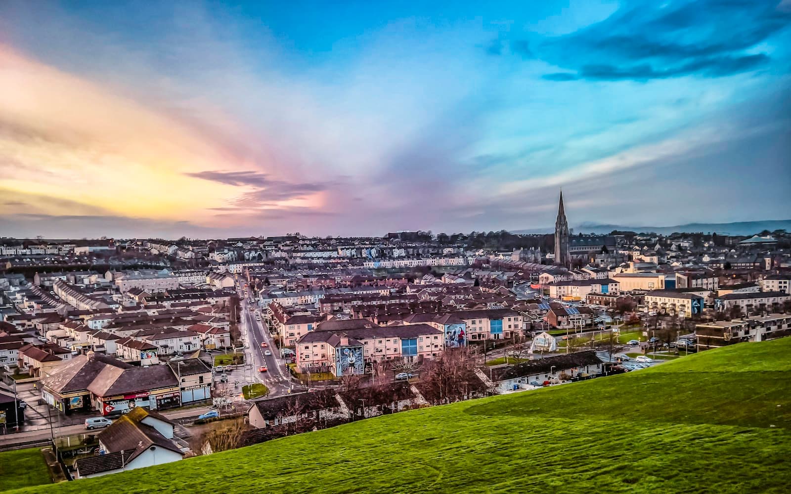 Aerial view of a town at sunset with houses, church spire, and dramatic purple-orange sky over green fields