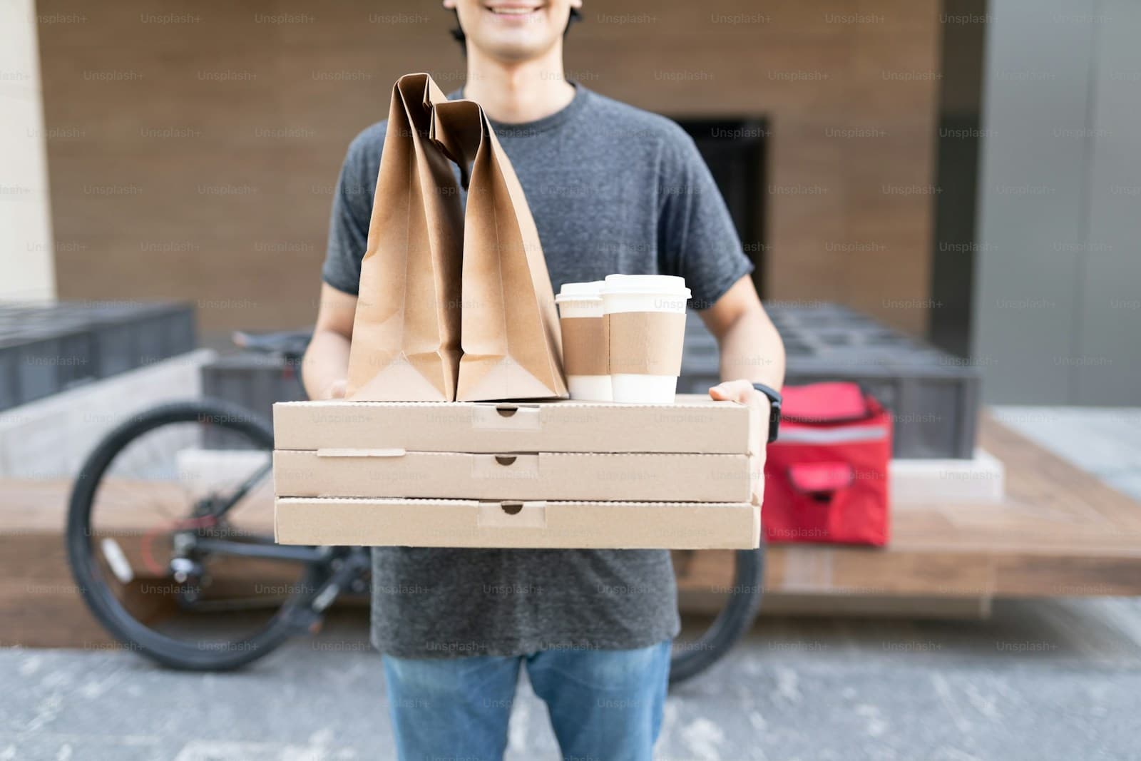 Smiling delivery rider holding pizza boxes beside a bicycle with a red insulated bag
