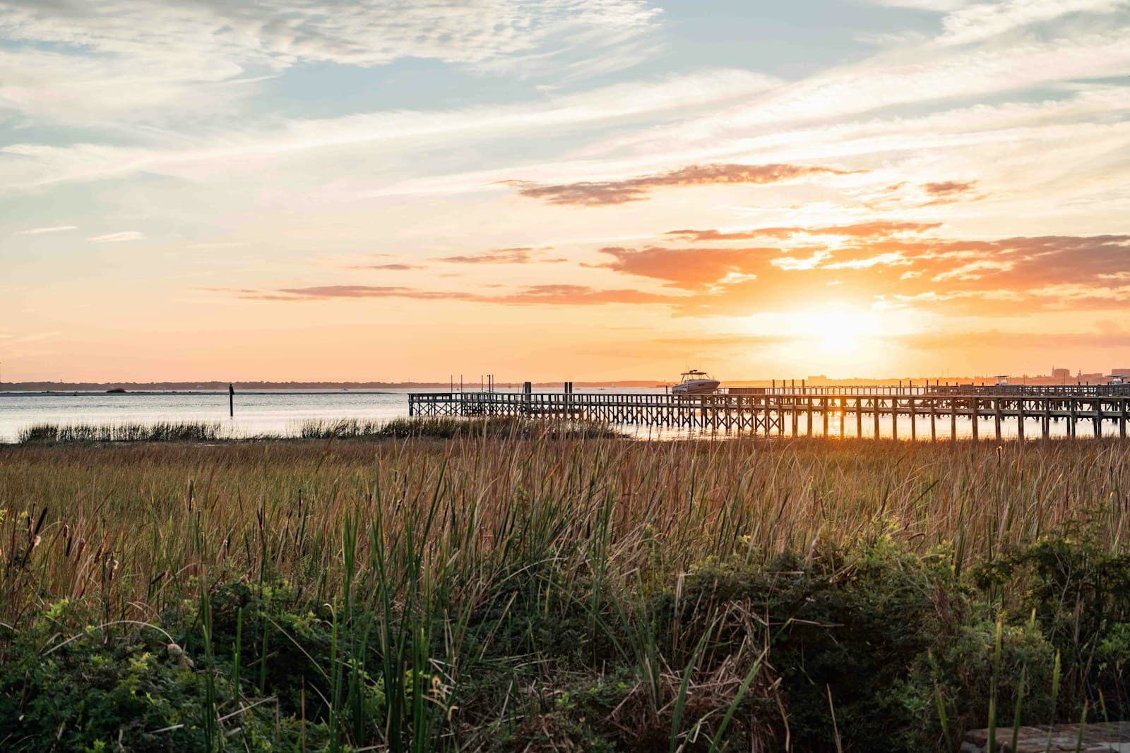 Wooden pier extending over water at sunset with marsh grasses in foreground and orange sky