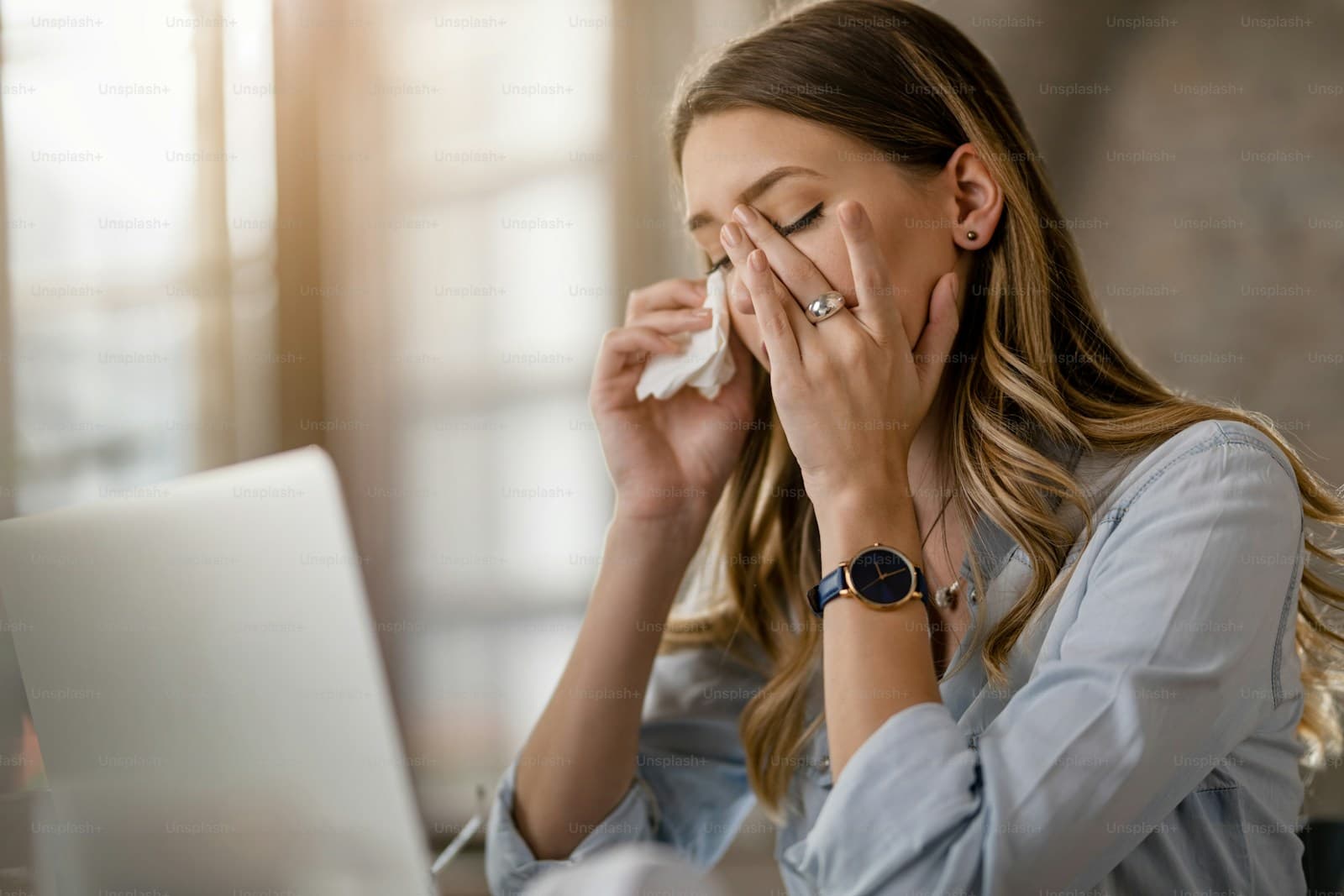Stressed woman covering face with hands while on phone call at desk with laptop in bright office setting