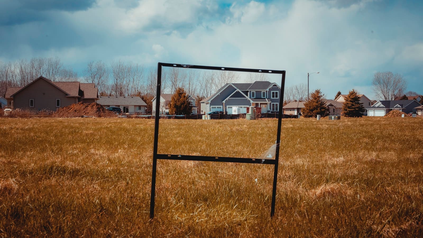 Empty metal sign frame in golden field with suburban houses in background under cloudy sky