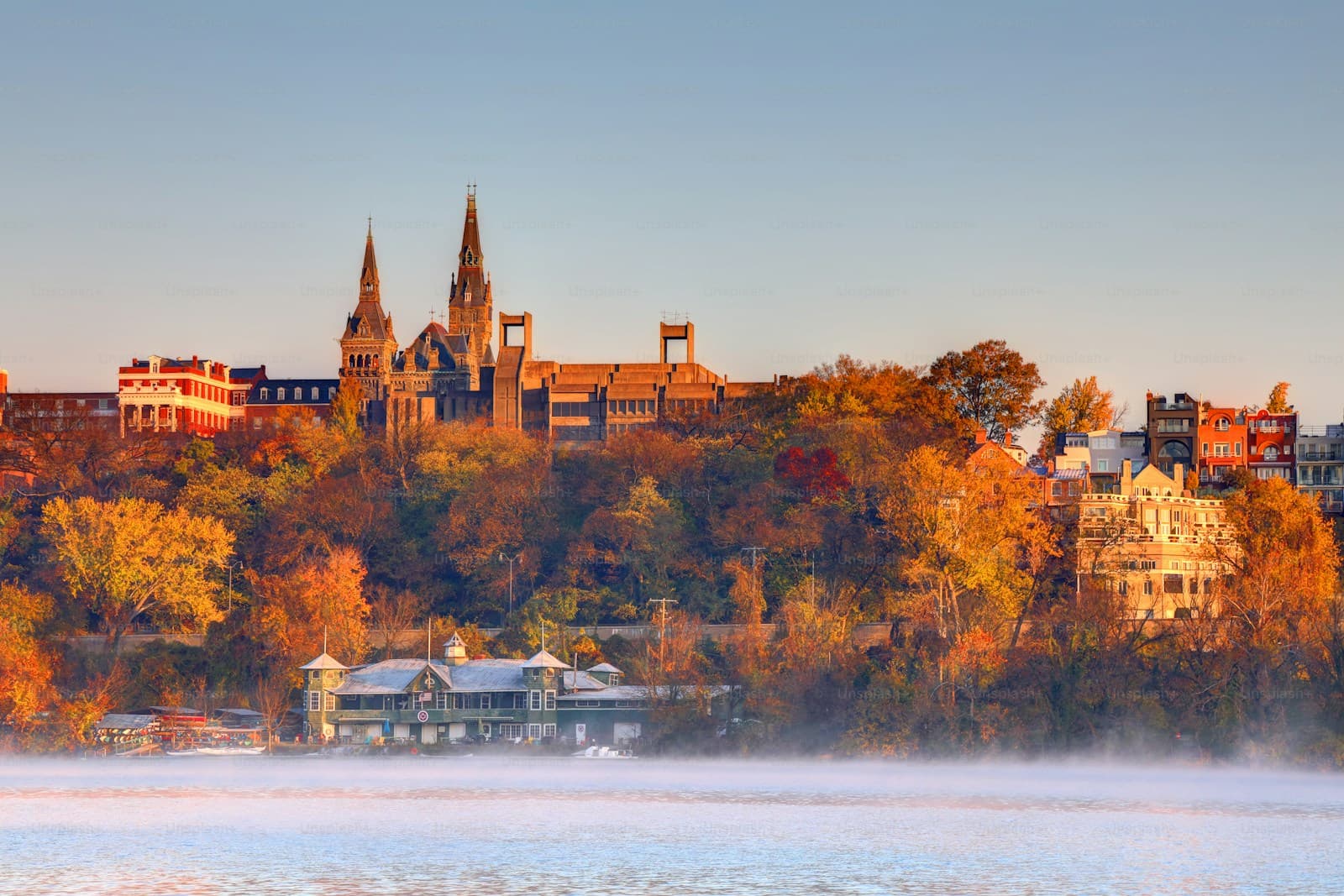Historic town with Gothic spires on hillside overlooking misty lake, surrounded by vibrant autumn foliage in golden light