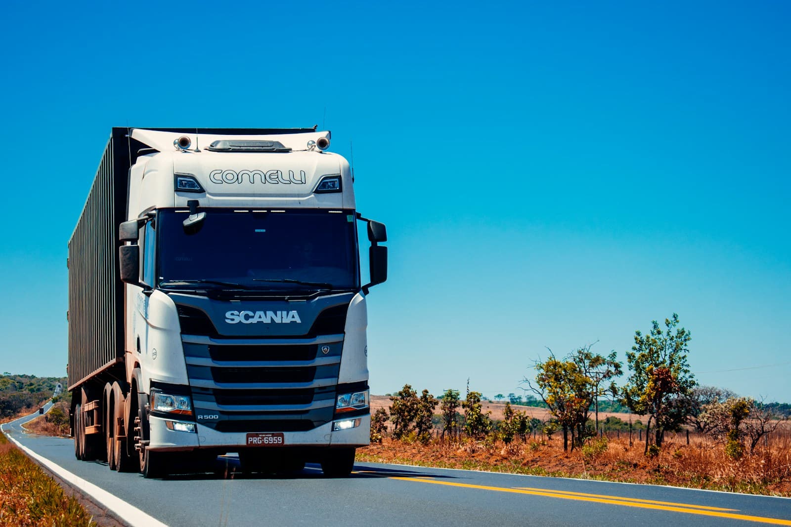 White Scania semi-truck and trailer on a rural highway amid autumn vegetation