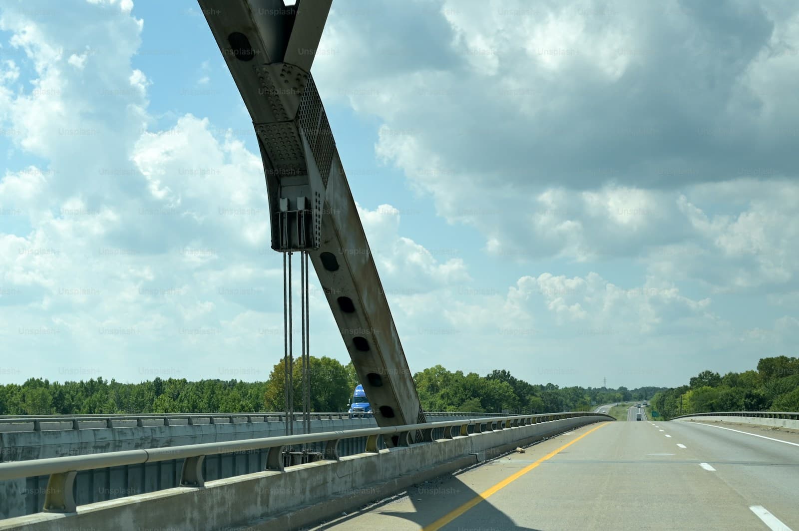 Highway bridge with concrete support beams and cables viewed from road with yellow lane markings under cloudy sky