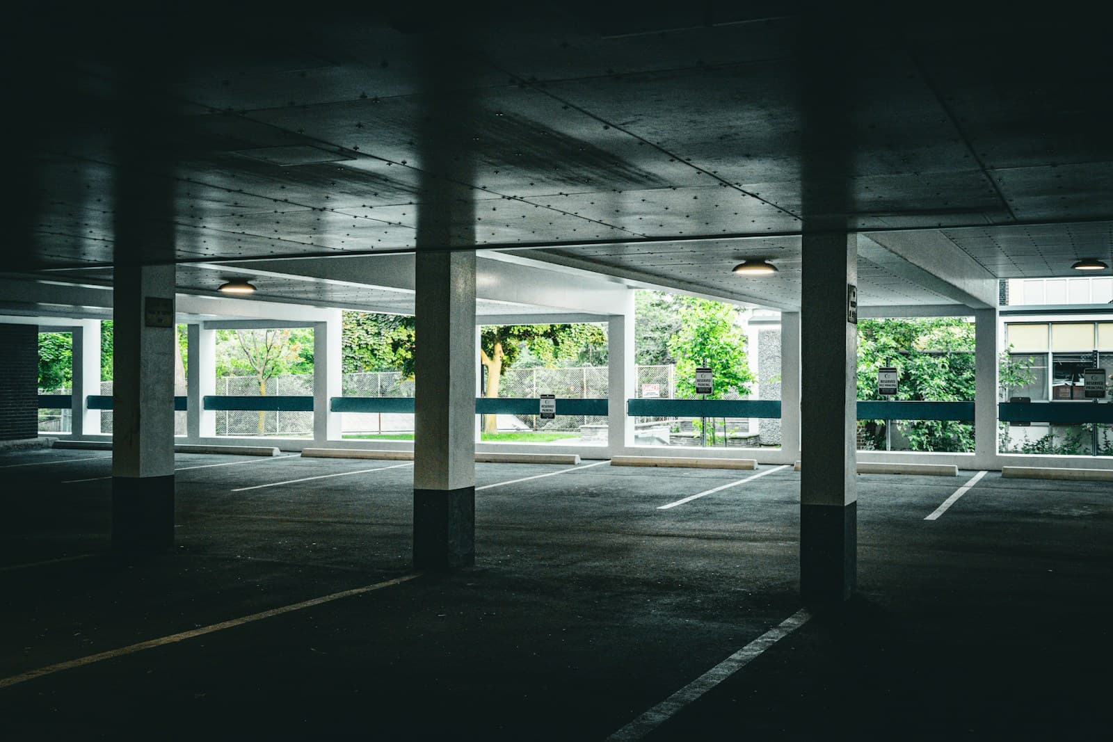 Empty concrete parking garage interior with fluorescent lights, pillars, and large windows showing trees outside