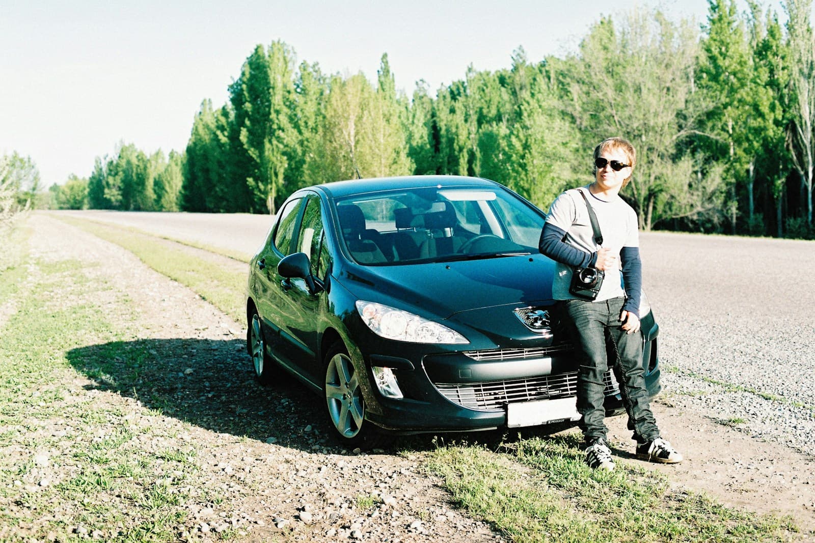 Person in sunglasses leaning against a black Peugeot parked on a sunny rural roadside