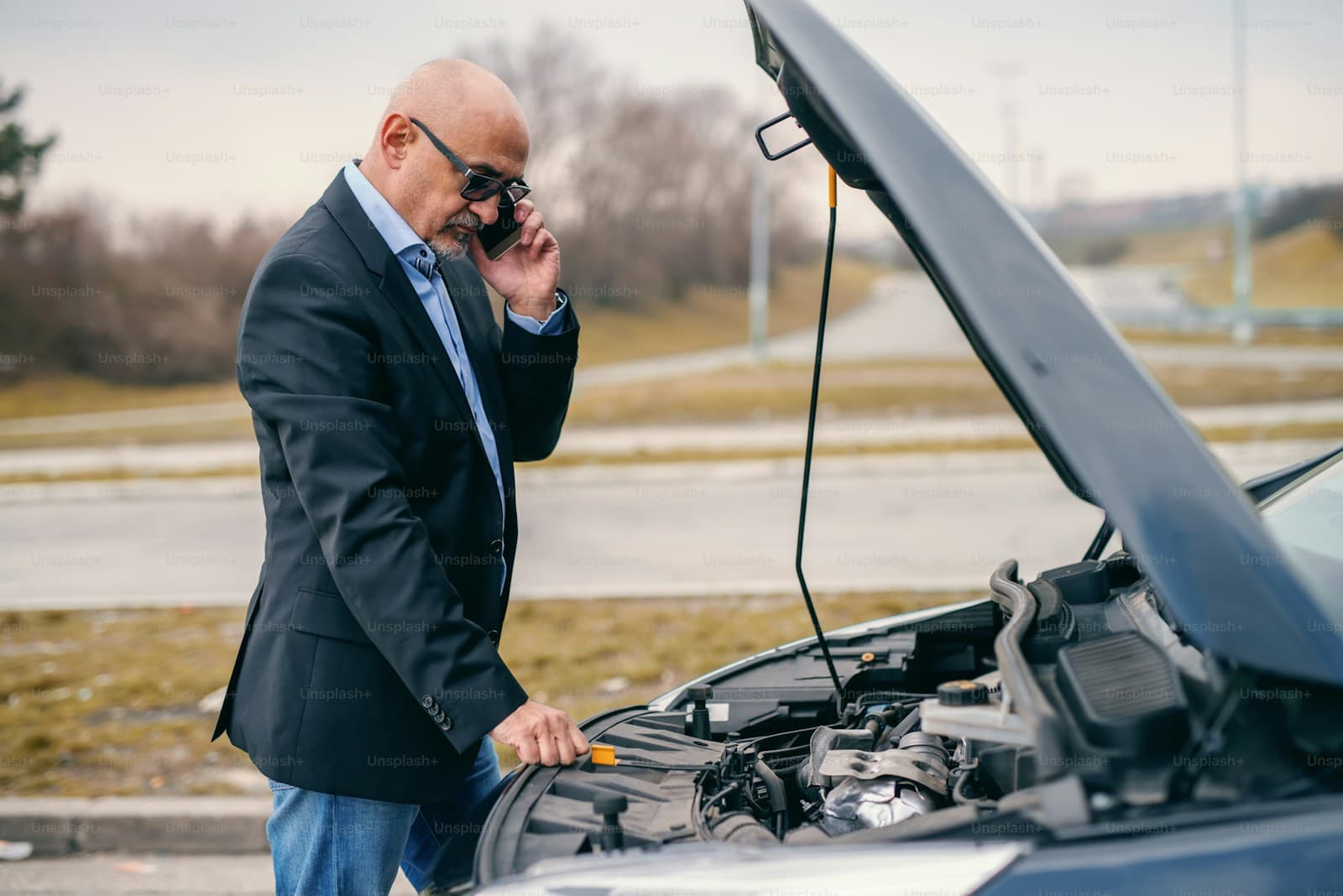 Man in a dark suit on his phone beside a car with the hood open after a breakdown