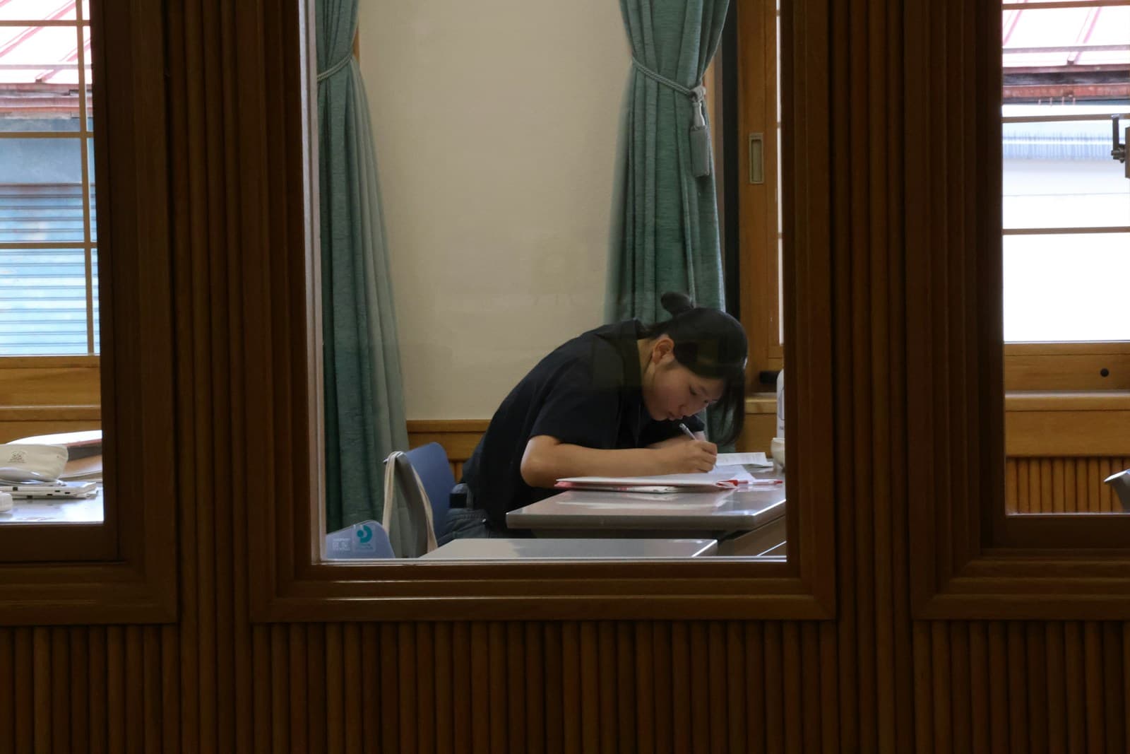 Person in dark clothing writing at desk viewed through window with wooden frame and curtains