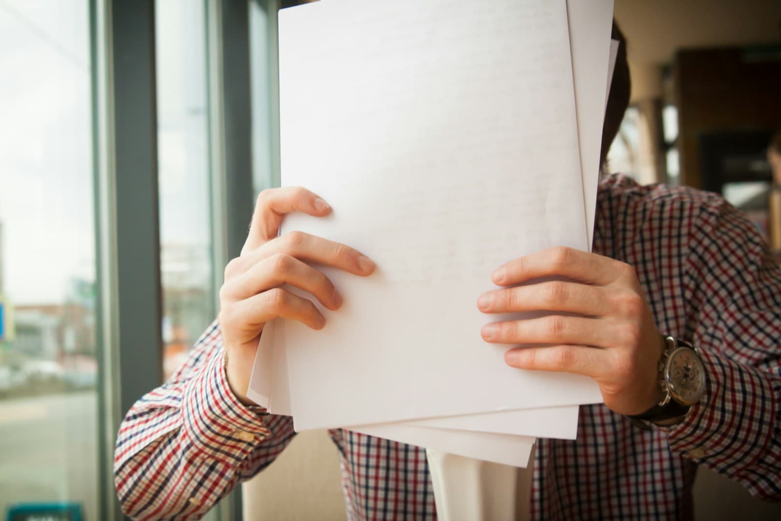 Person in plaid shirt holding blank white paper document near office window