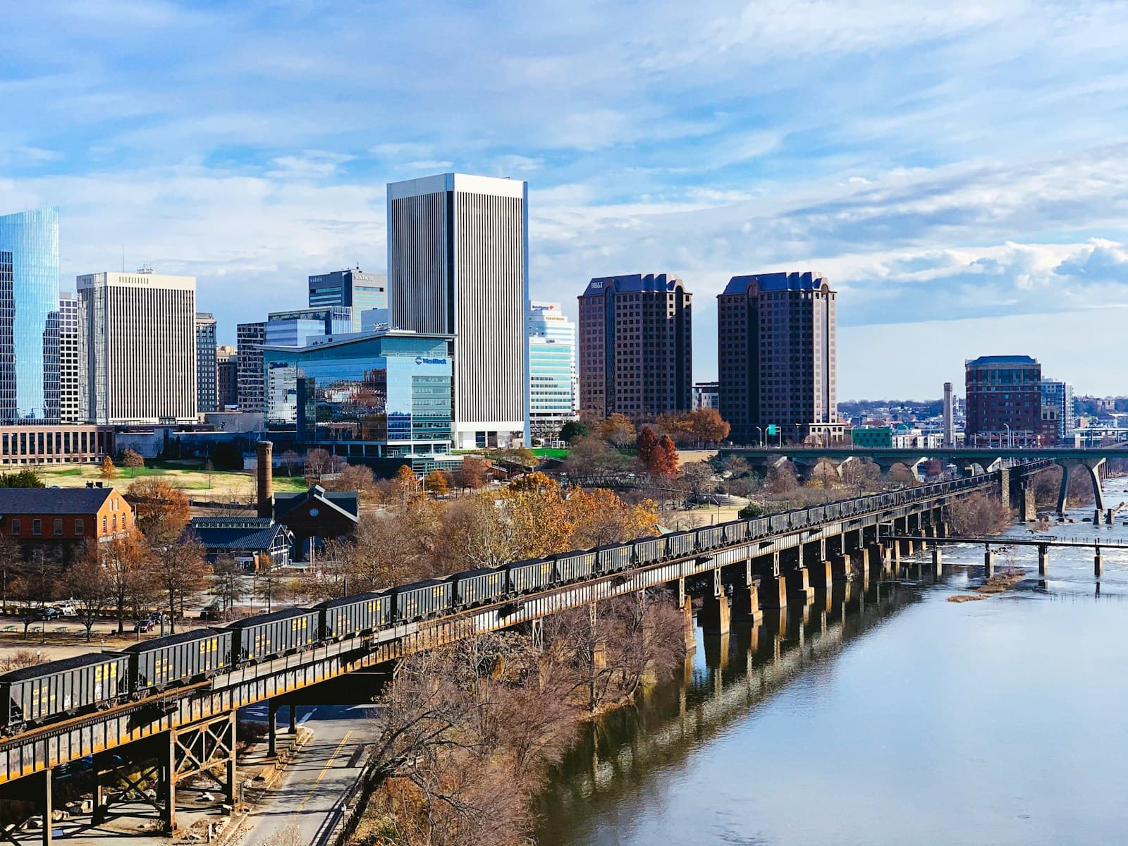 City skyline with modern buildings and pedestrian bridge crossing a river, surrounded by autumn trees