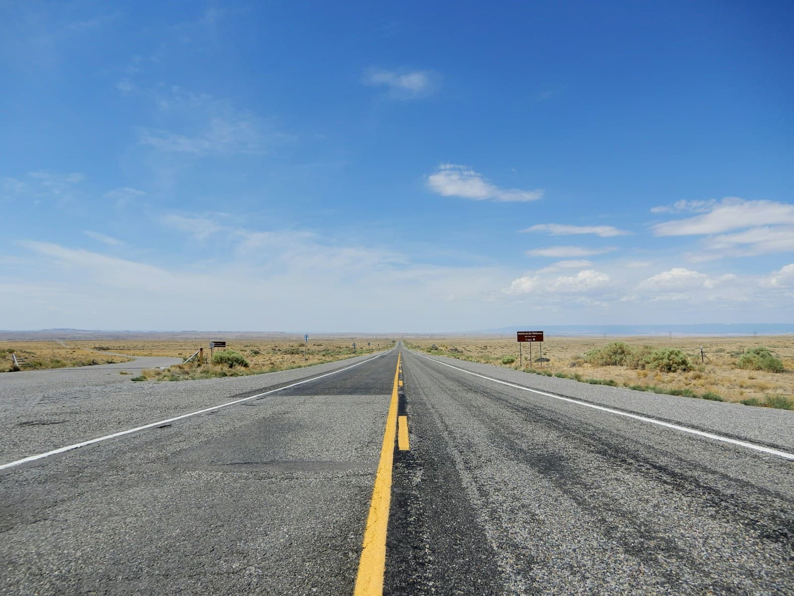 Straight desert highway with yellow center line stretching to horizon under blue sky with scattered clouds