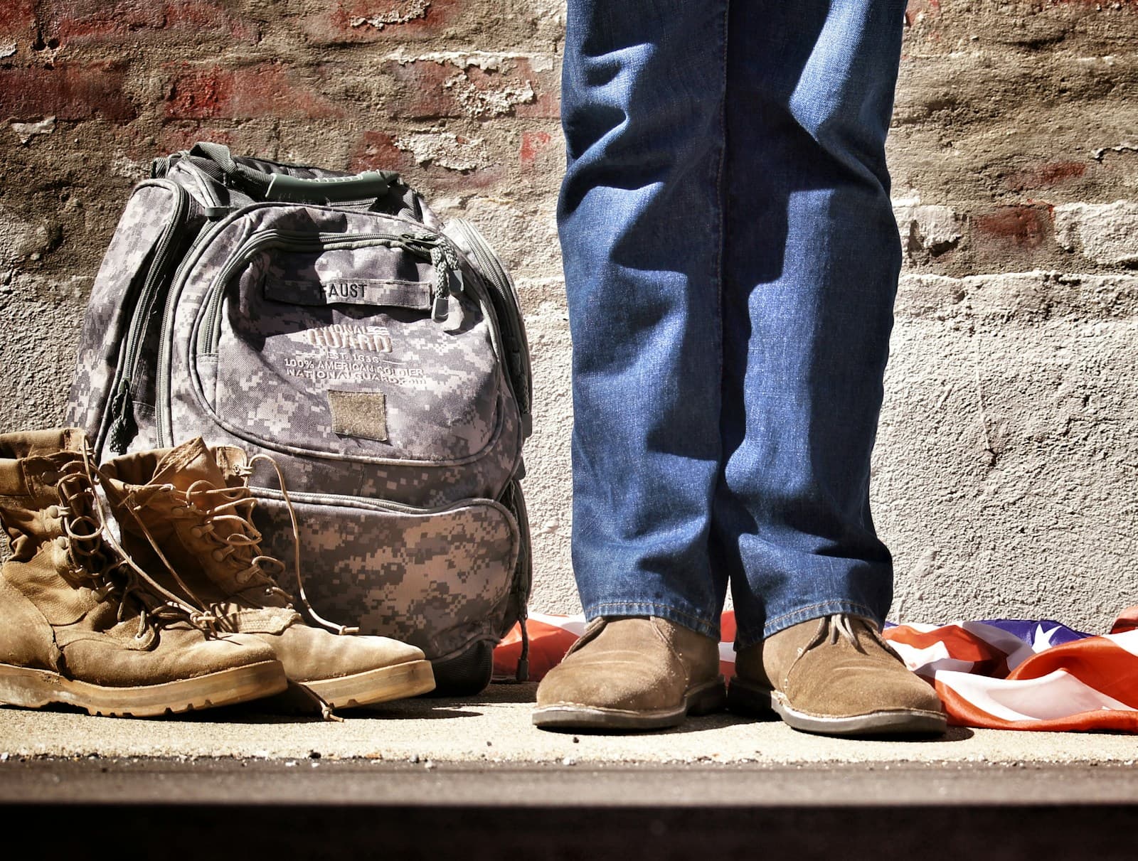 Person in jeans and work boots beside a military pack, combat boots and a folded flag