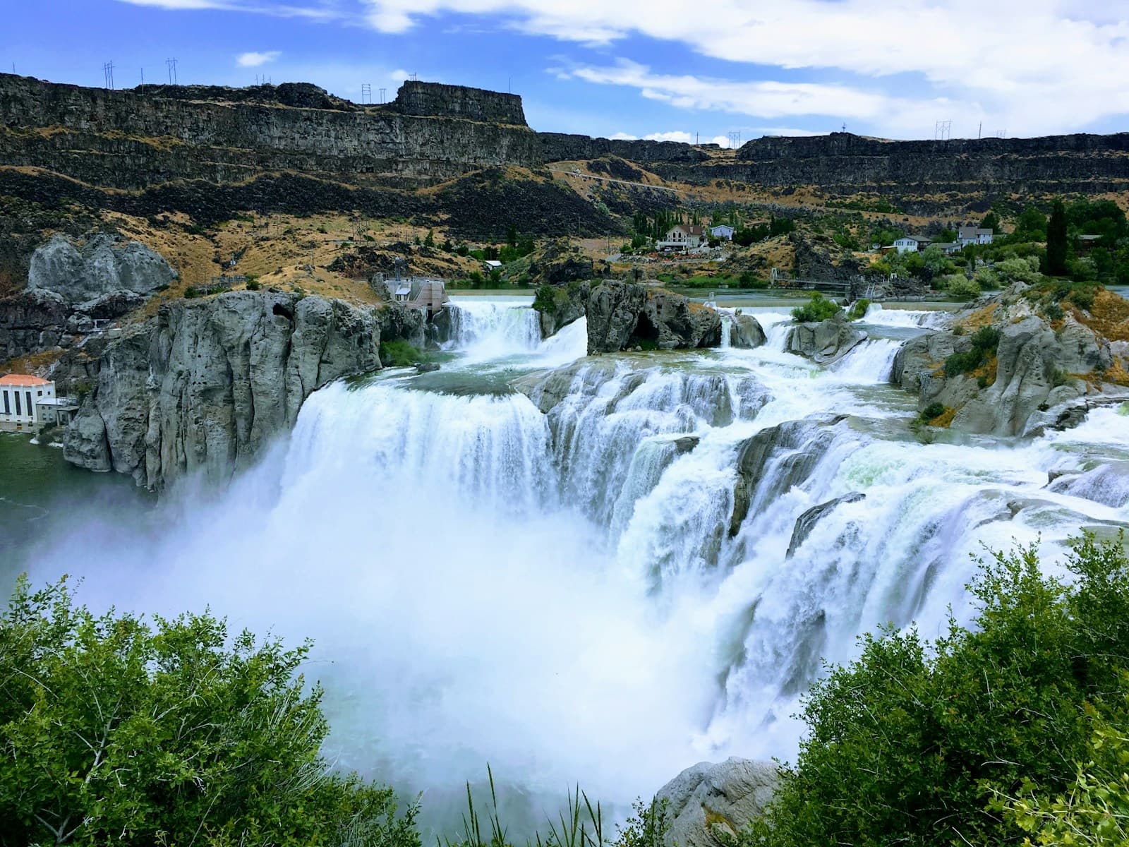 Multi-tiered waterfall flowing over dark rocky cliffs in a desert canyon with green vegetation and blue sky