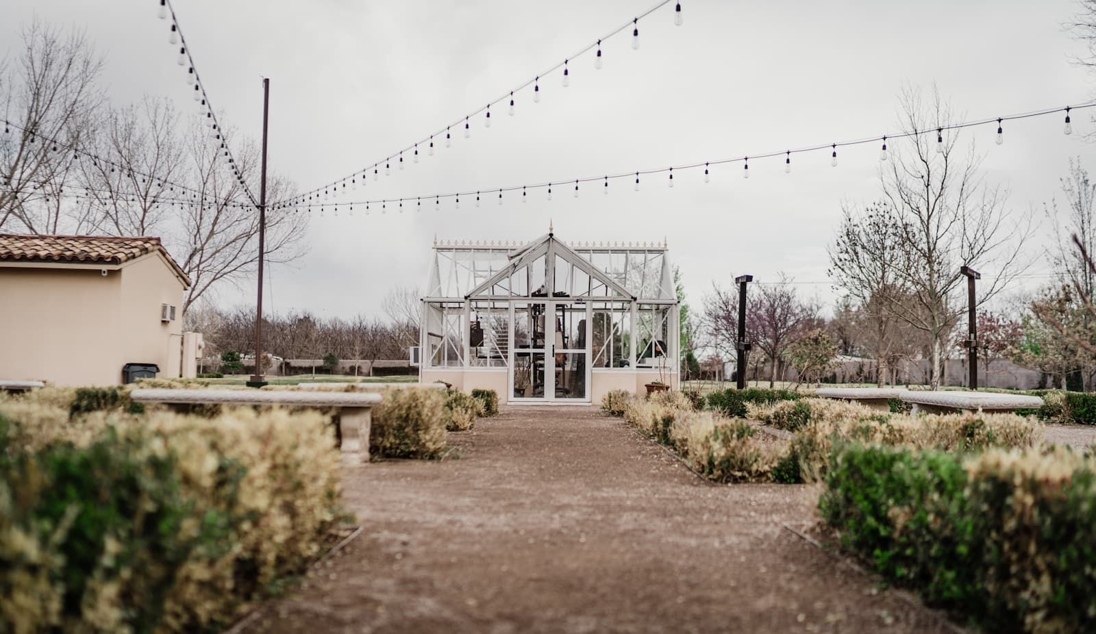 White greenhouse pavilion wedding venue with string lights and landscaped pathways under overcast sky