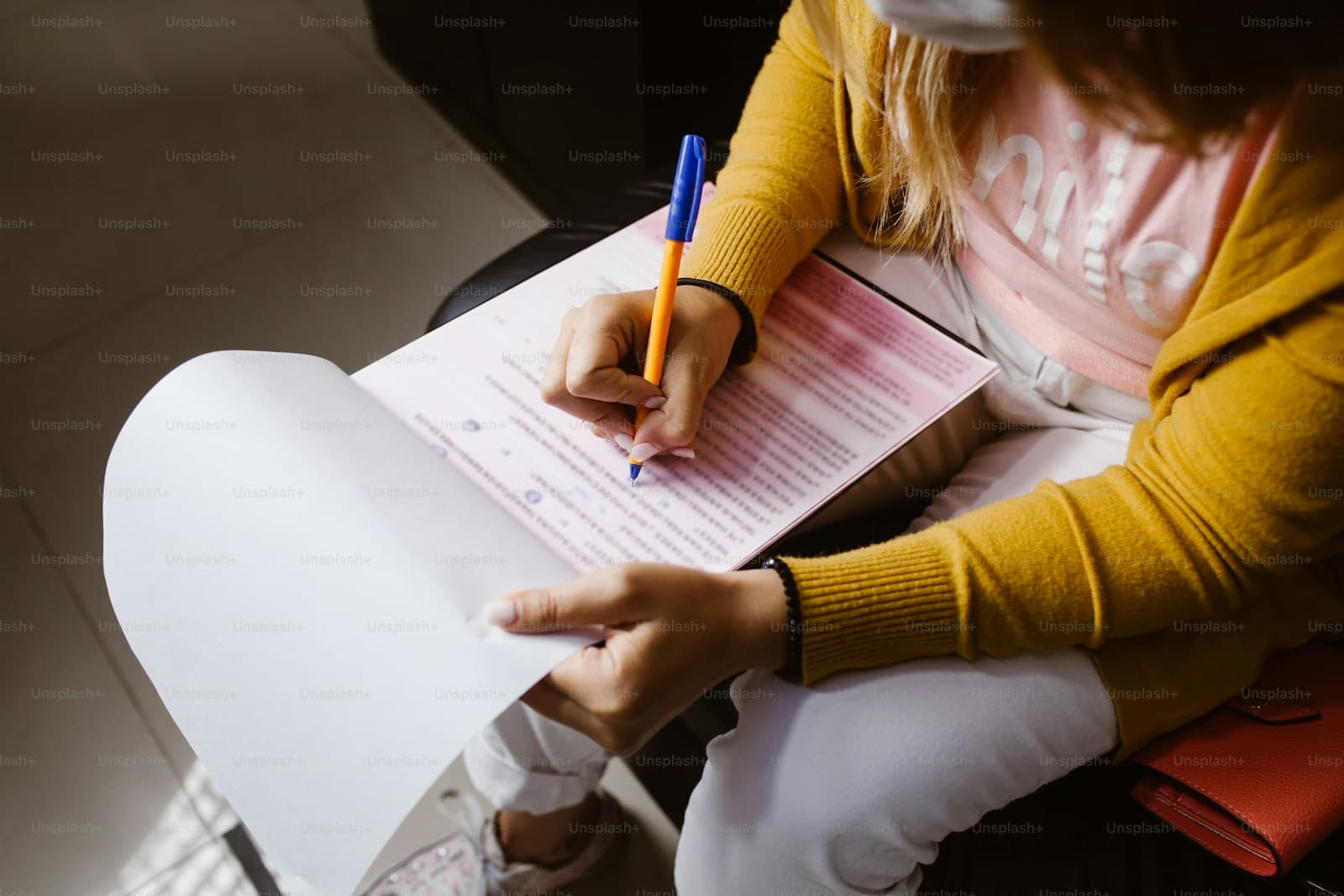 Person in yellow sweater sitting cross-legged writing on a form or document with a blue pen