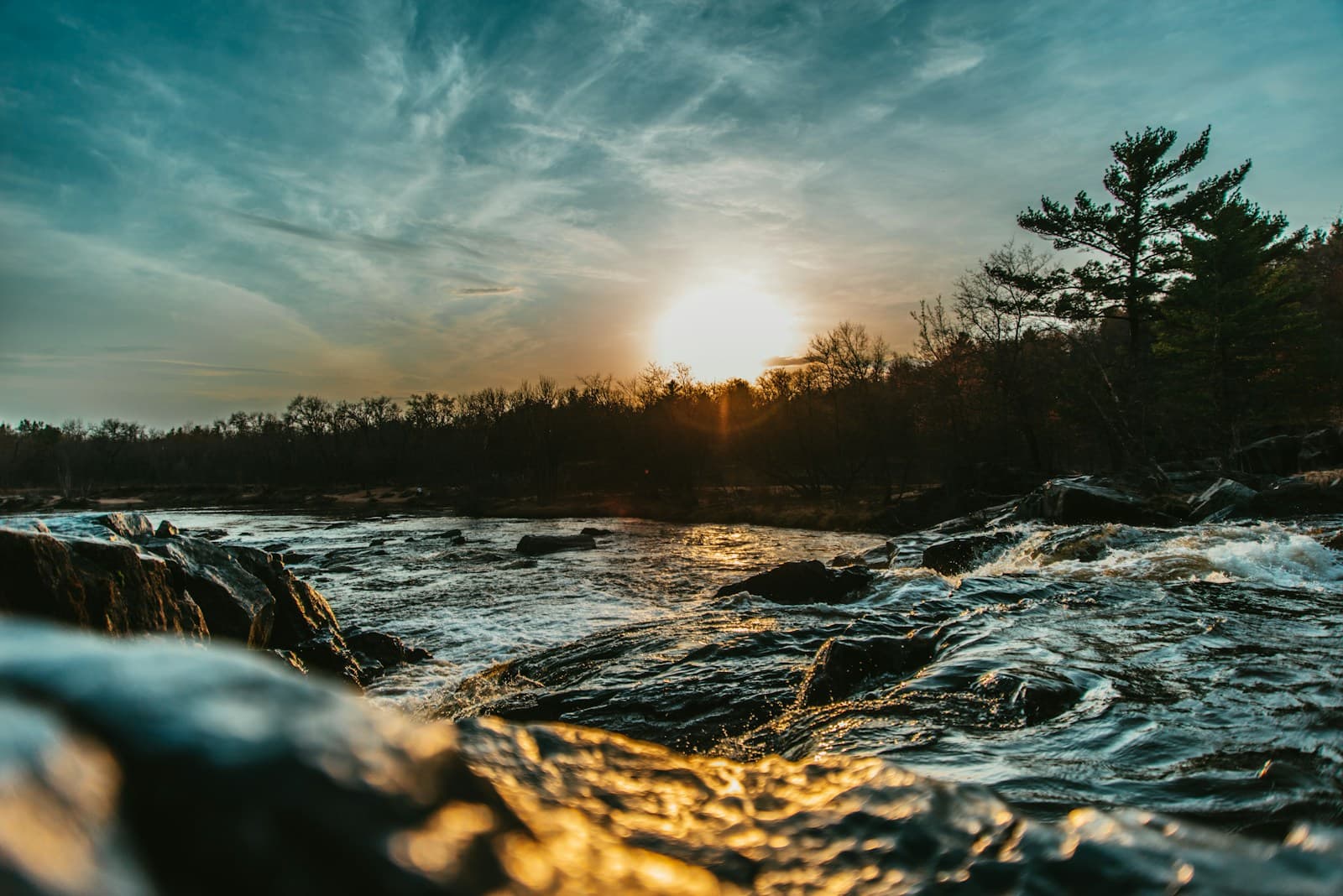 Sunset over a flowing river with rocky terrain and trees silhouetted against a dramatic cloudy sky