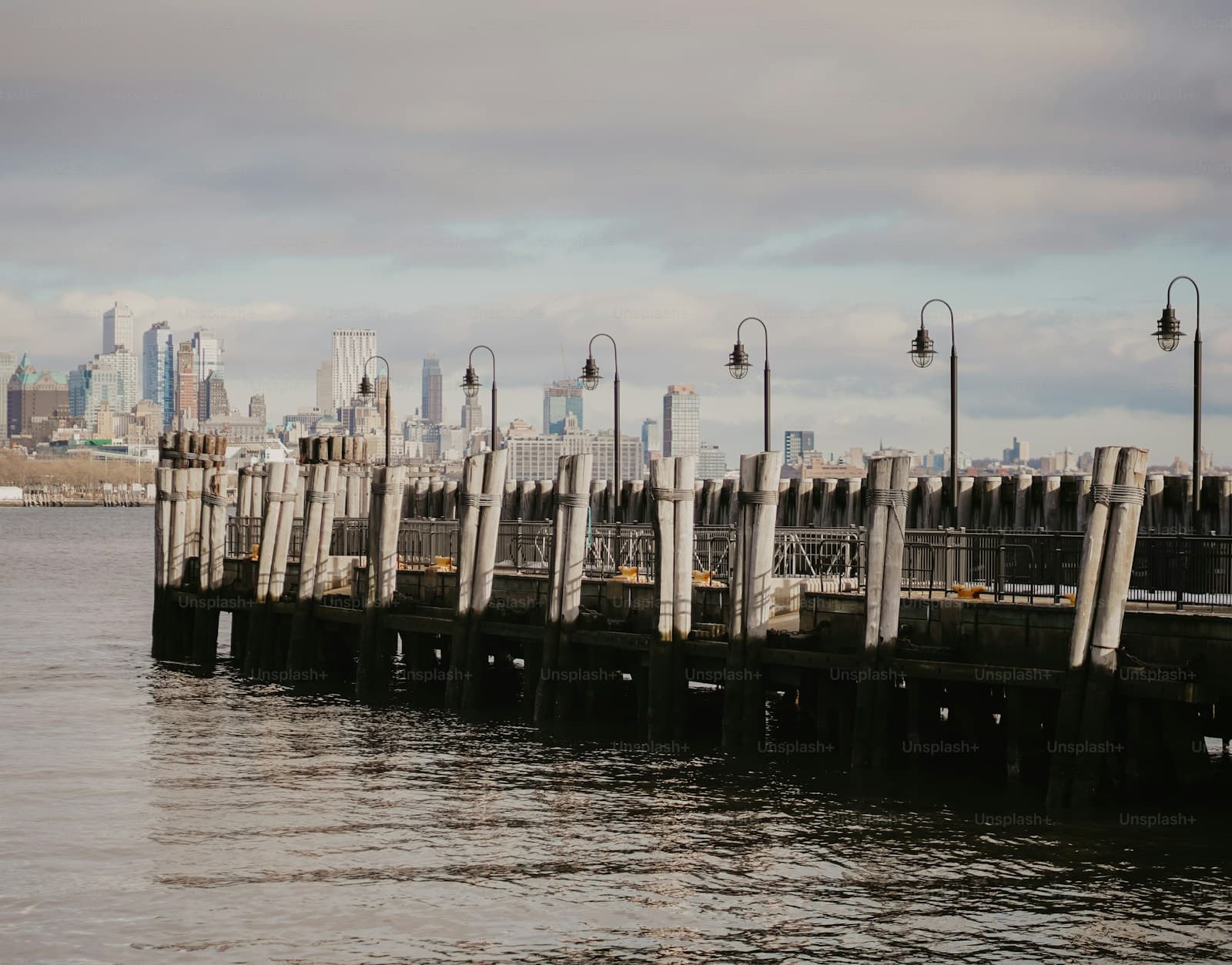 Wooden pier with lamp posts extending into water with city skyline in background under cloudy sky