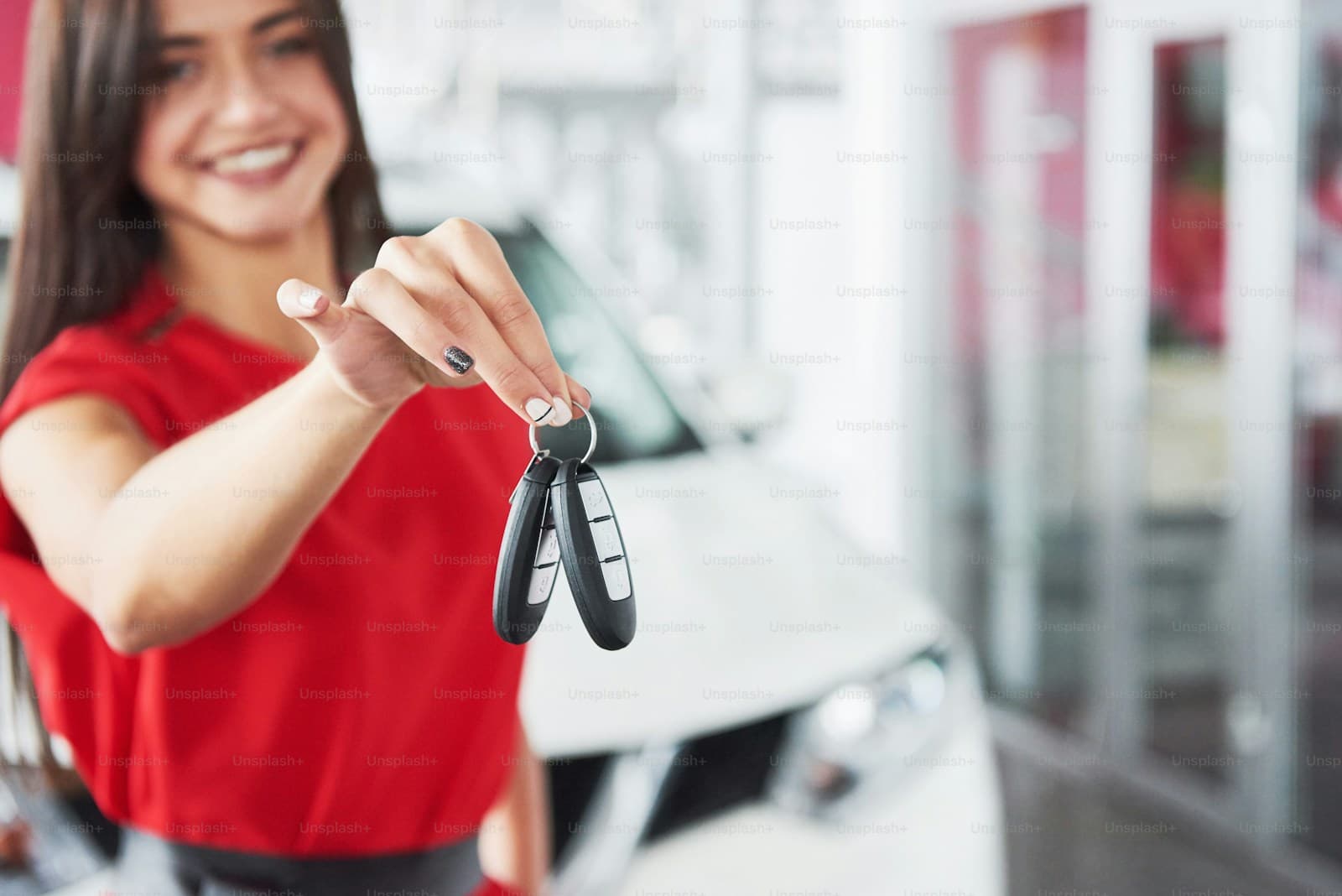 Smiling young woman in a red shirt holding out car keys at a dealership lot