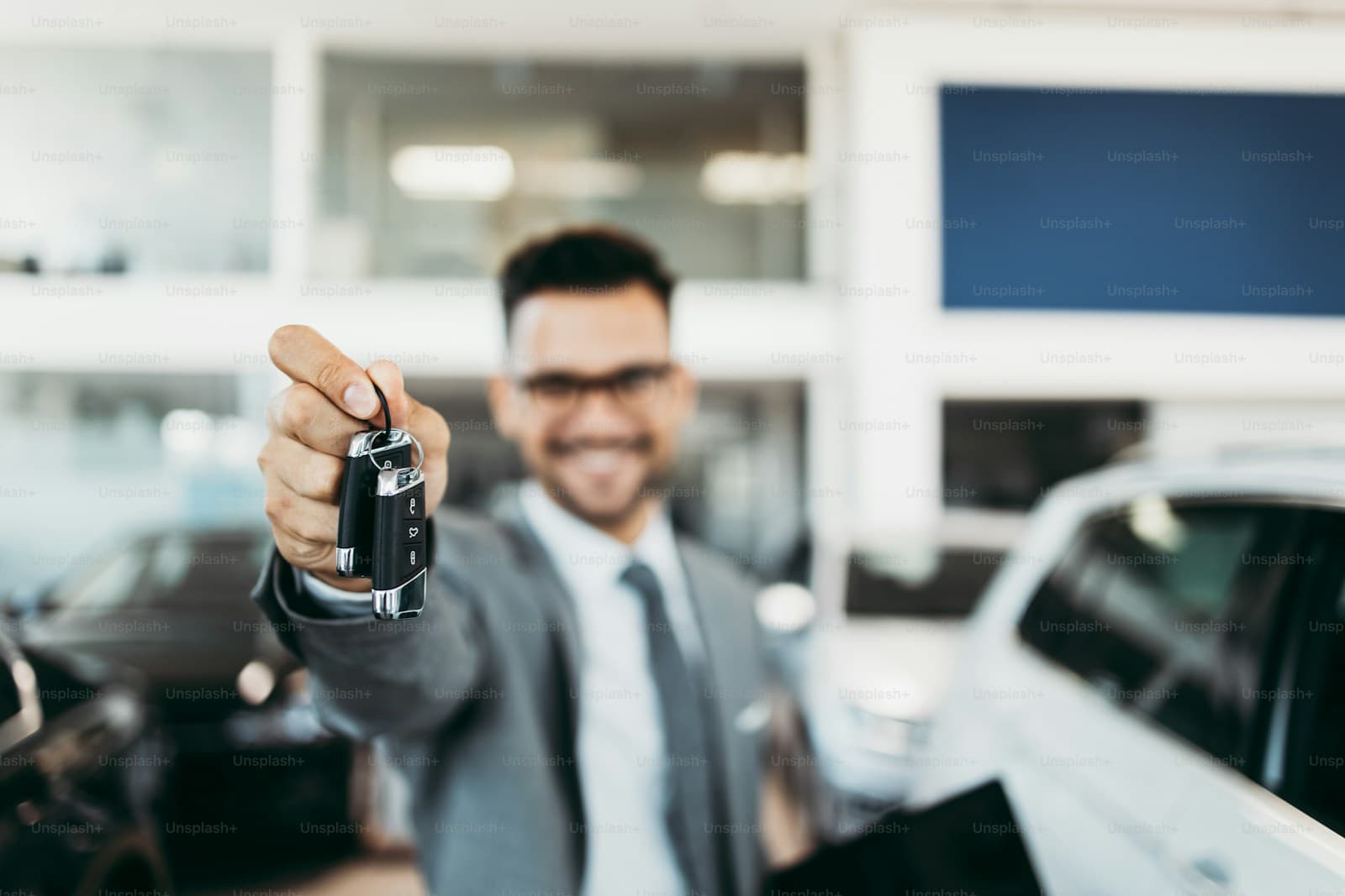 Smiling salesman in a suit holding out car keys in a dealership showroom