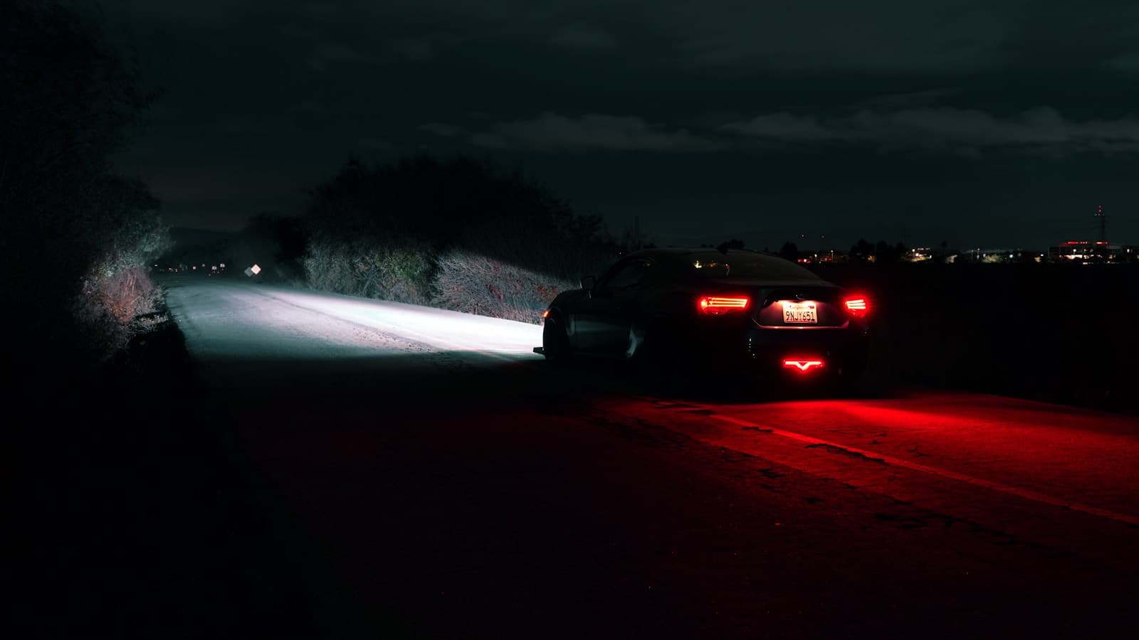 Two cars on dark road at night with bright headlights and red taillights illuminating the pavement