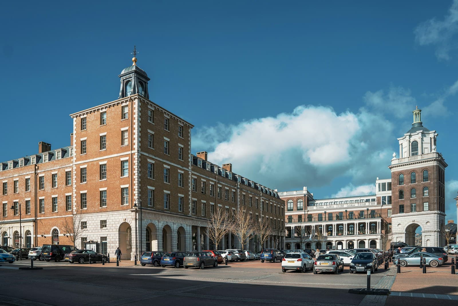 Large red brick institutional buildings with white trim and cupolas arranged around a courtyard with parked cars