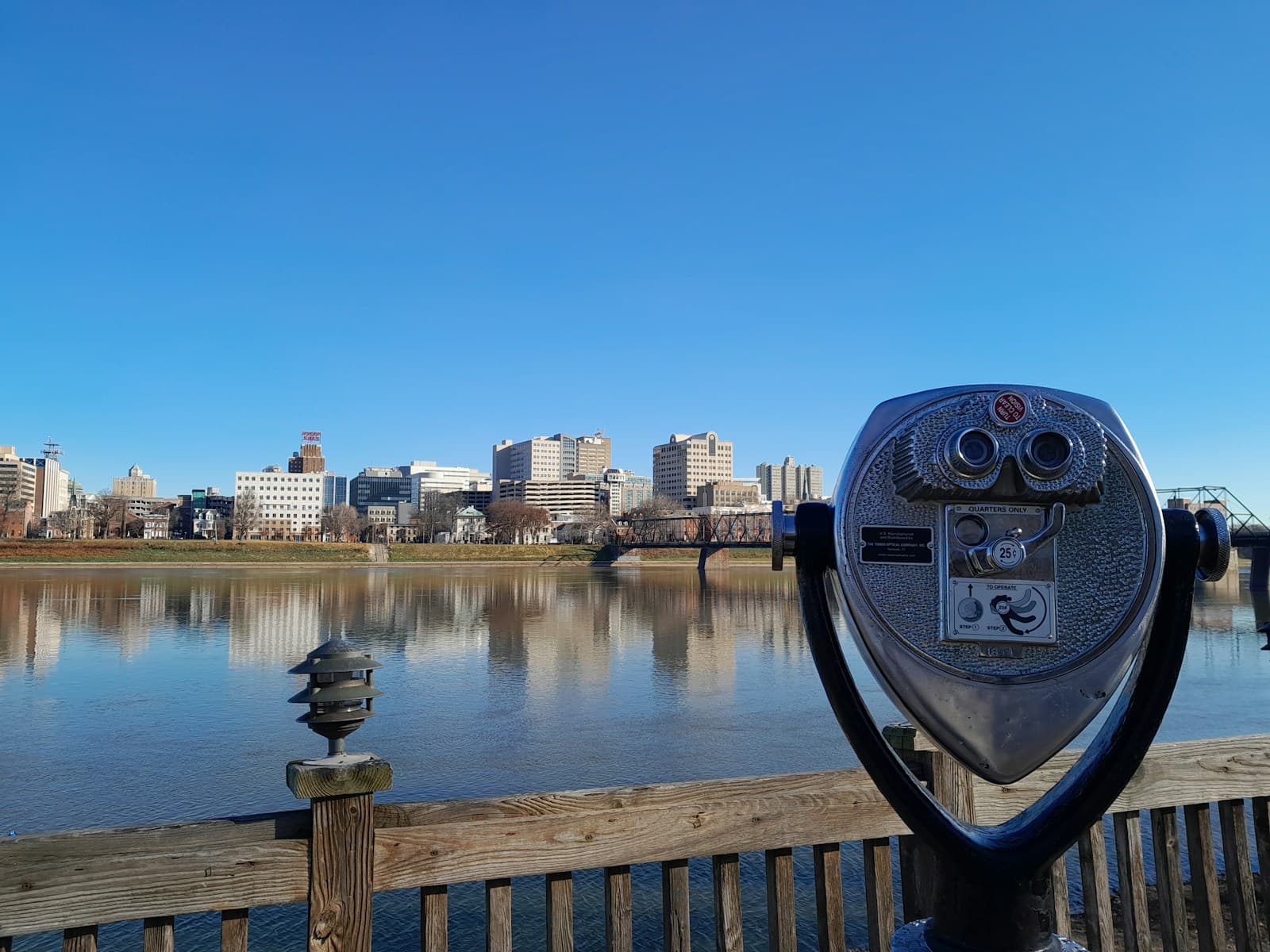 Coin-operated telescope overlooking city skyline reflected in calm water from wooden boardwalk