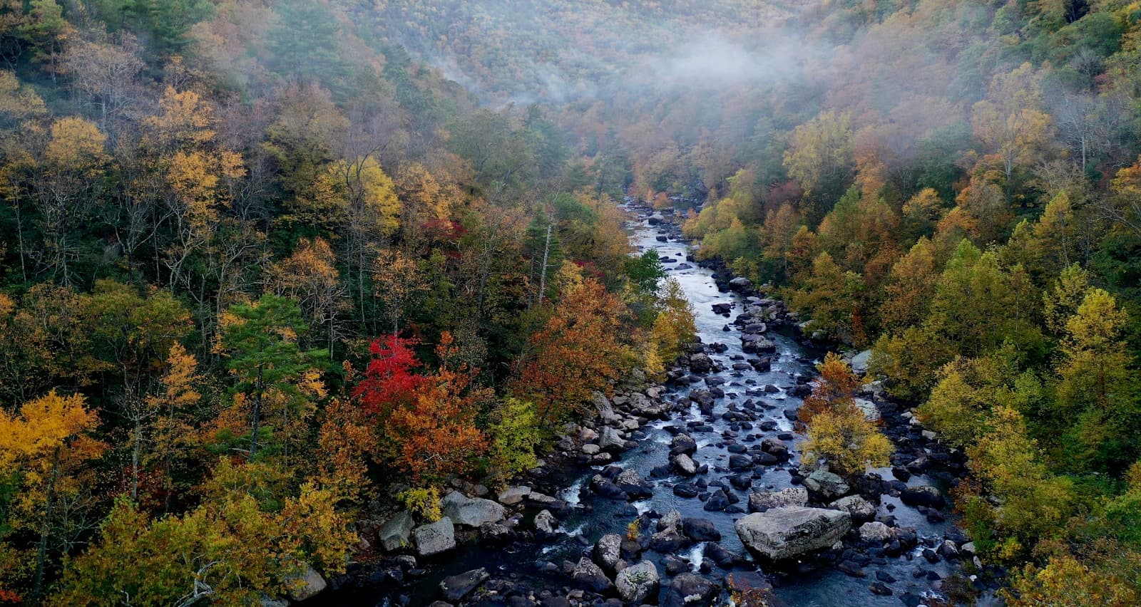 Aerial view of a river flowing through autumn forest with colorful fall foliage and misty hills