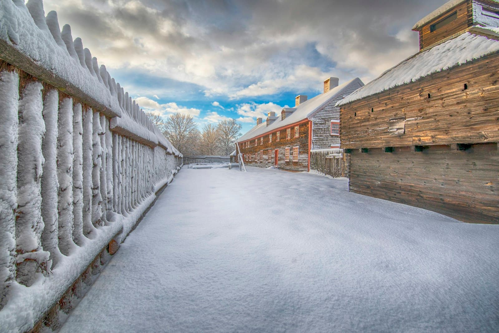 Snow-covered wooden walkway with white railings between rustic log buildings in winter village setting
