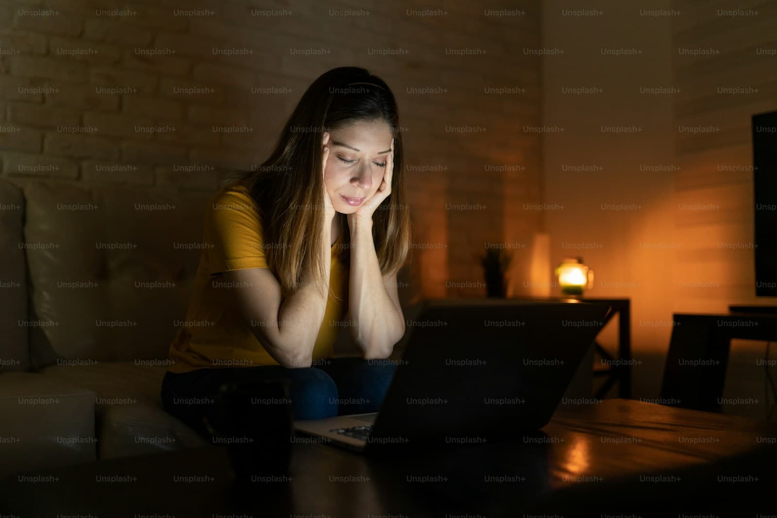 Woman working late on laptop computer in dimly lit room, looking tired with chin resting on hands