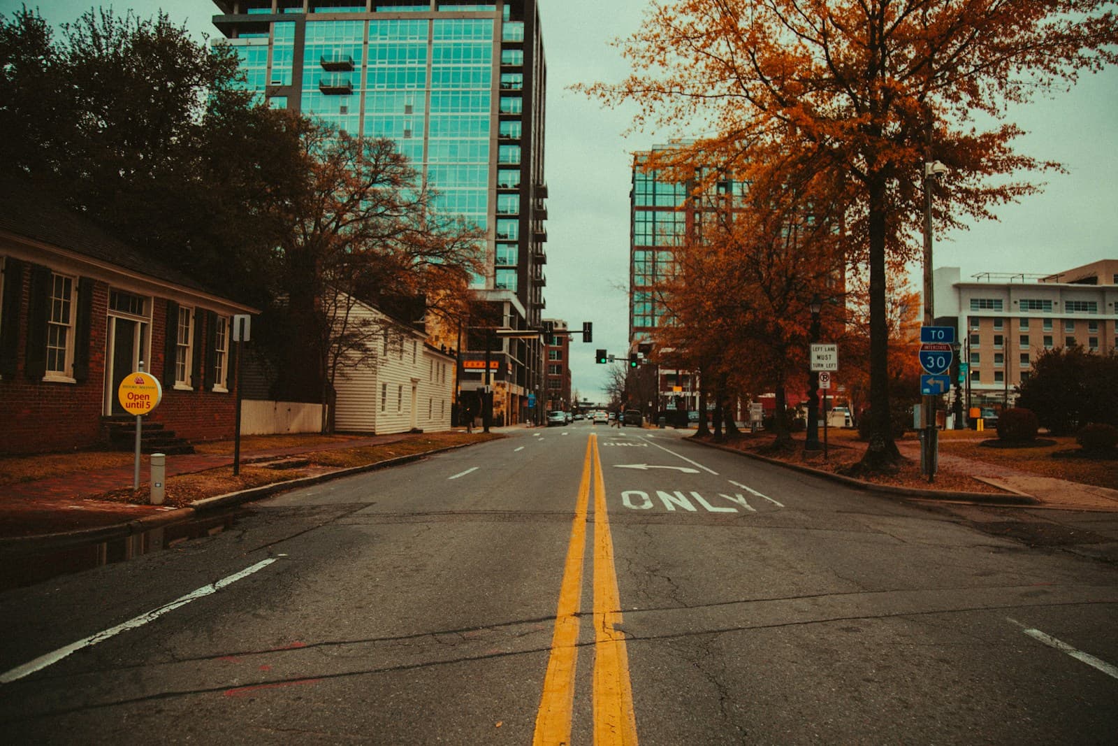 Urban street with yellow lane markings leading to glass buildings, lined with autumn trees in warm vintage tones