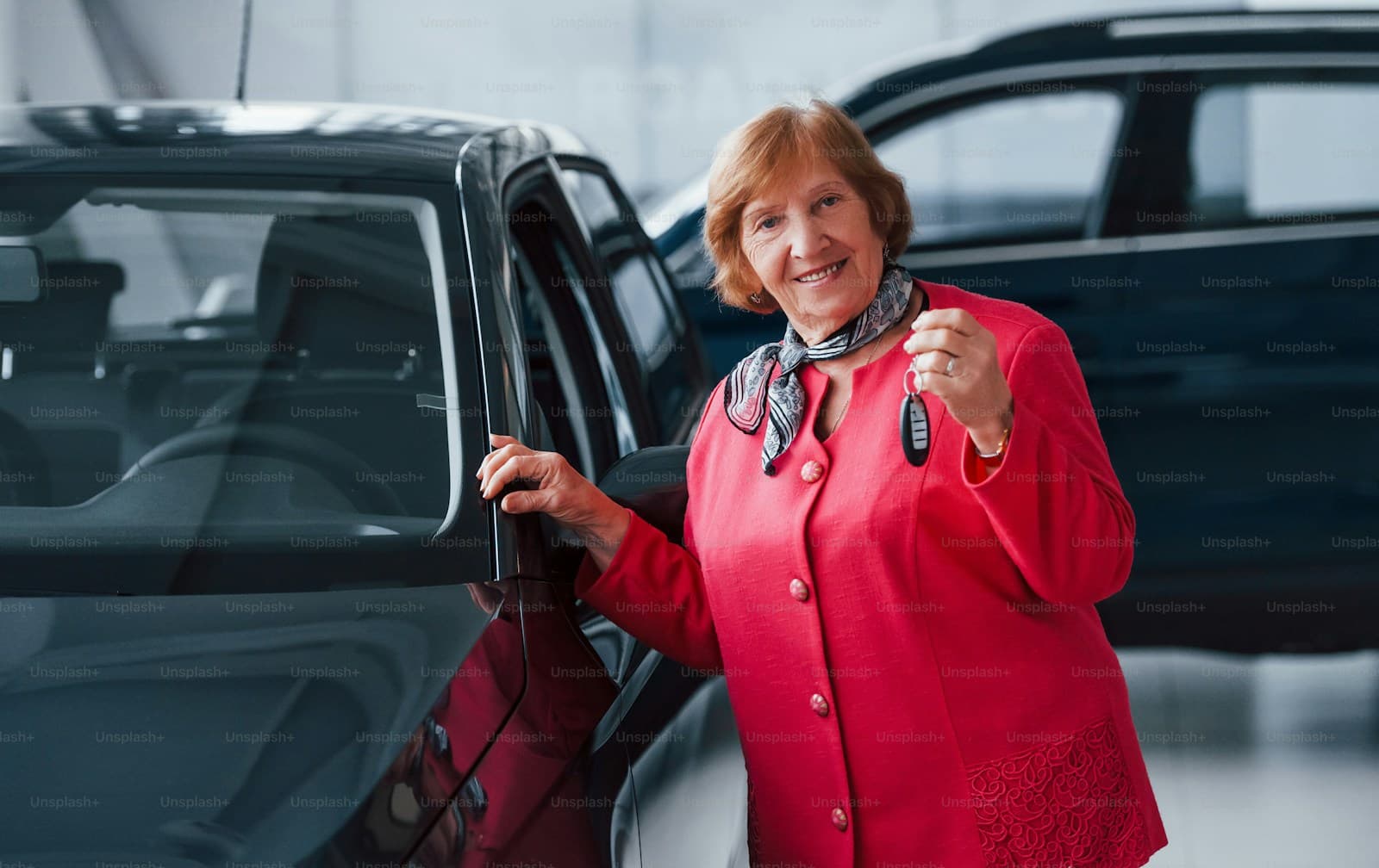 Happy woman in red coat holding car keys next to new dark car in dealership showroom