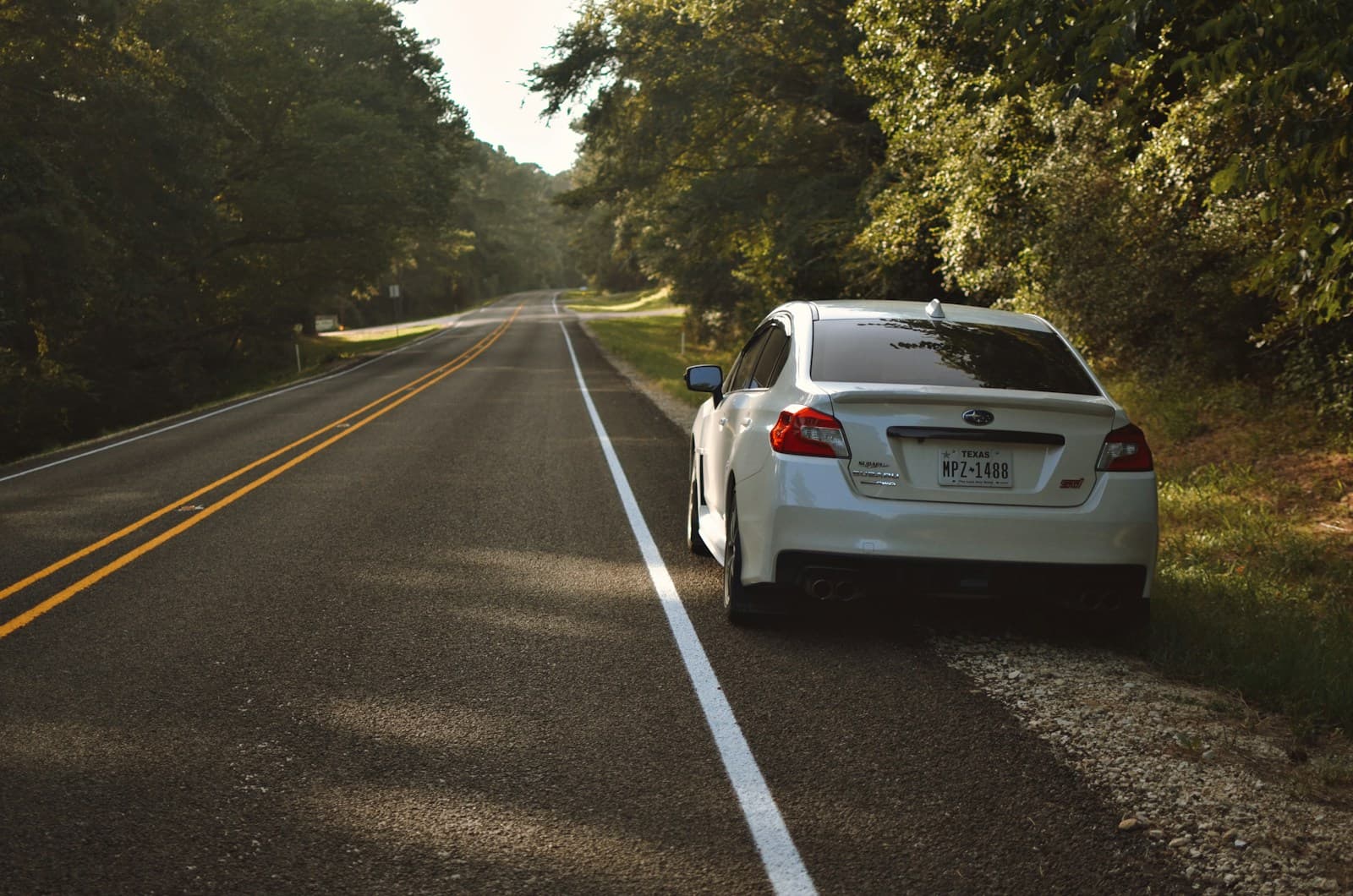 White sedan pulled onto the shoulder of a tree-lined rural highway in late afternoon light