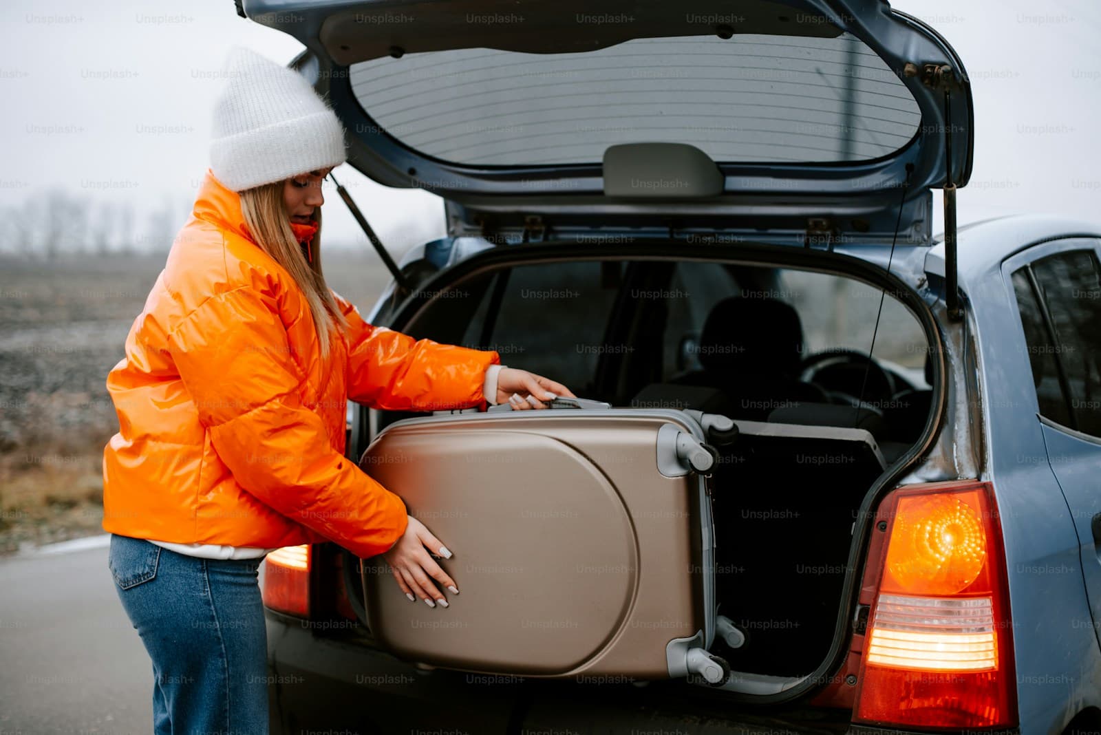 Traveler in an orange jacket loading a suitcase into an SUV trunk on a rural road