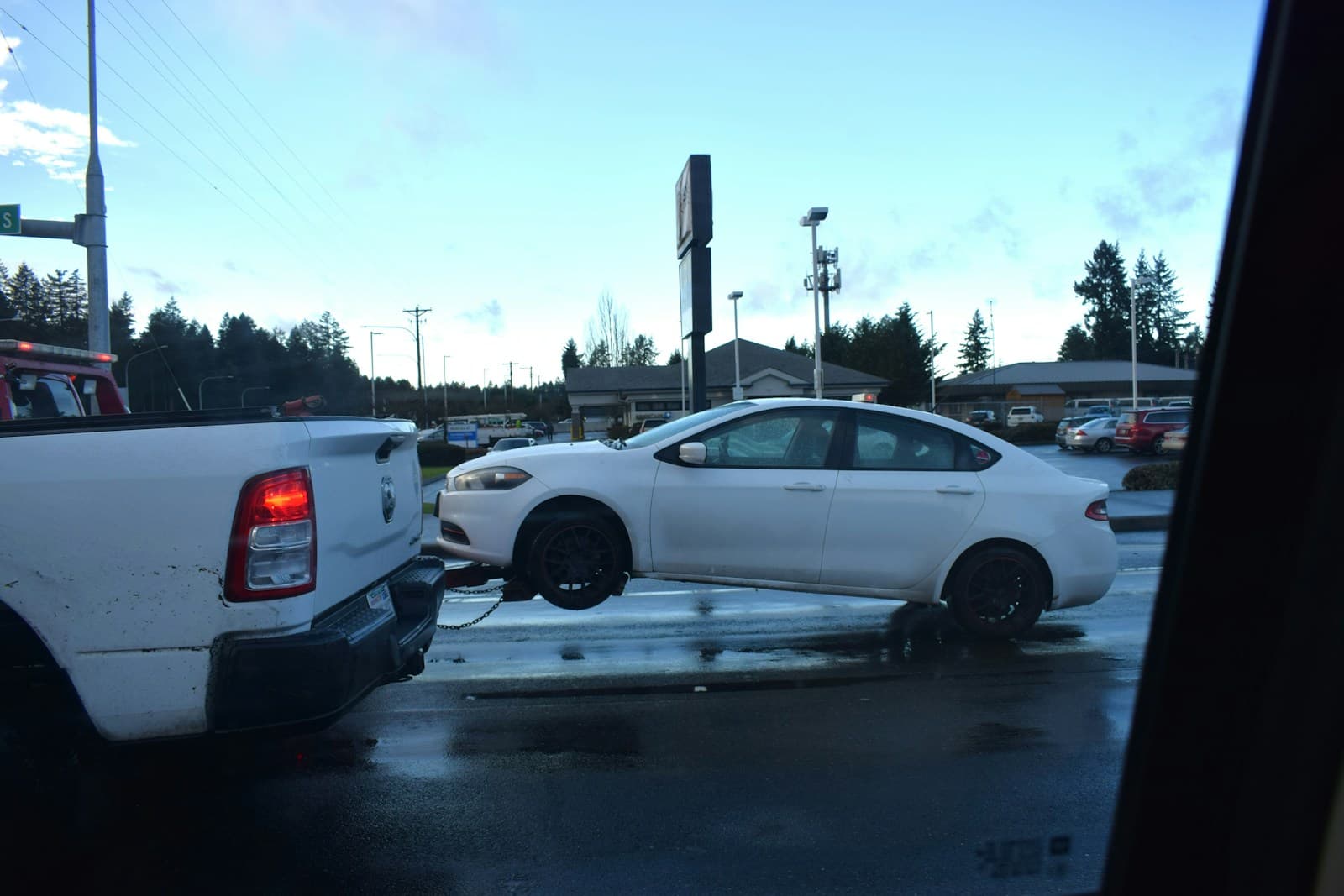 White pickup towing a sedan on a flatbed trailer across a lot on an overcast day