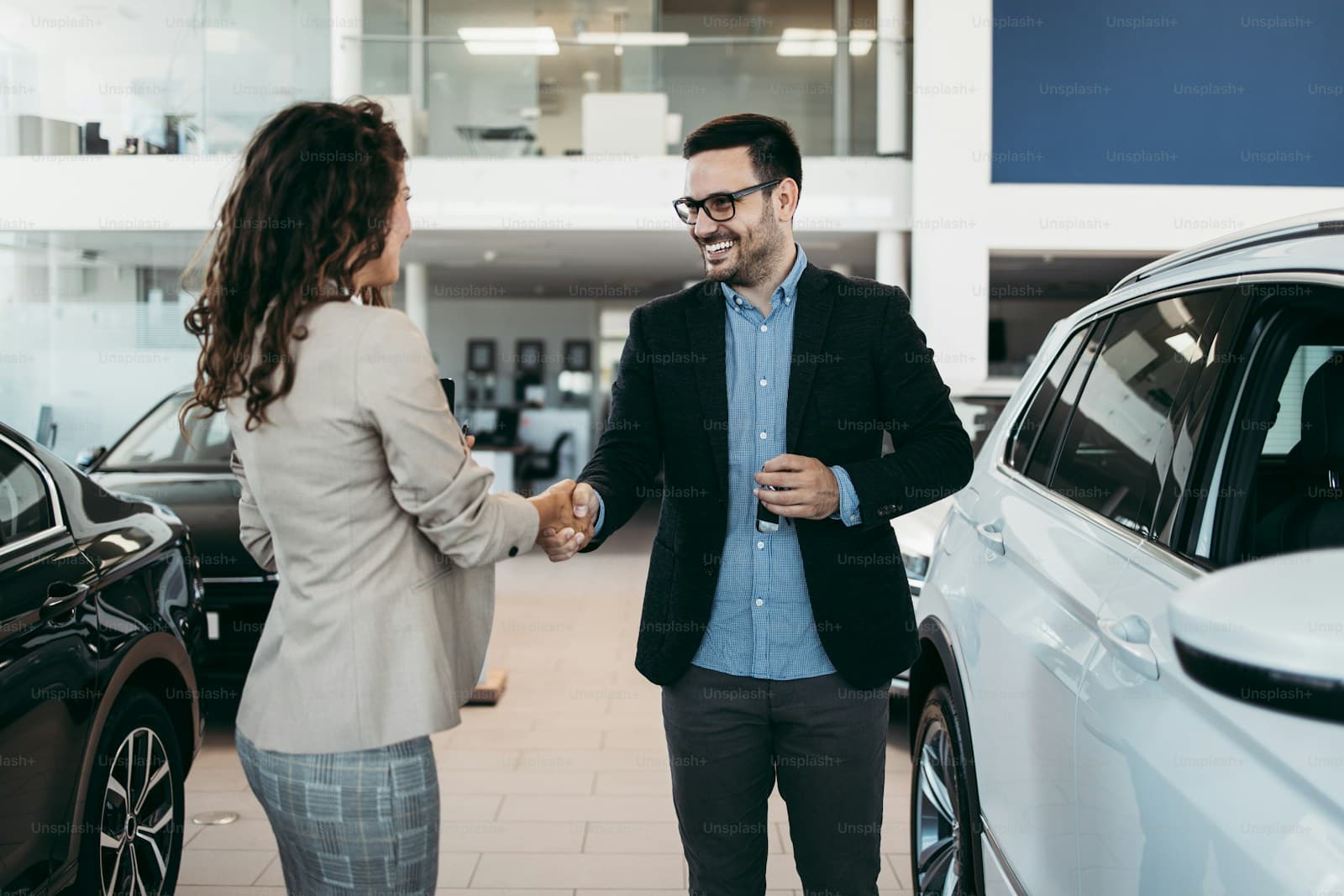 Woman in a beige blazer shaking hands with a salesman in a dealership showroom