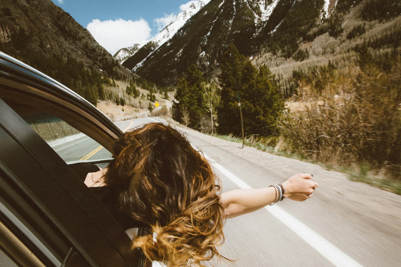 Person with flowing hair leaning out car window on scenic mountain road with snow-capped peaks