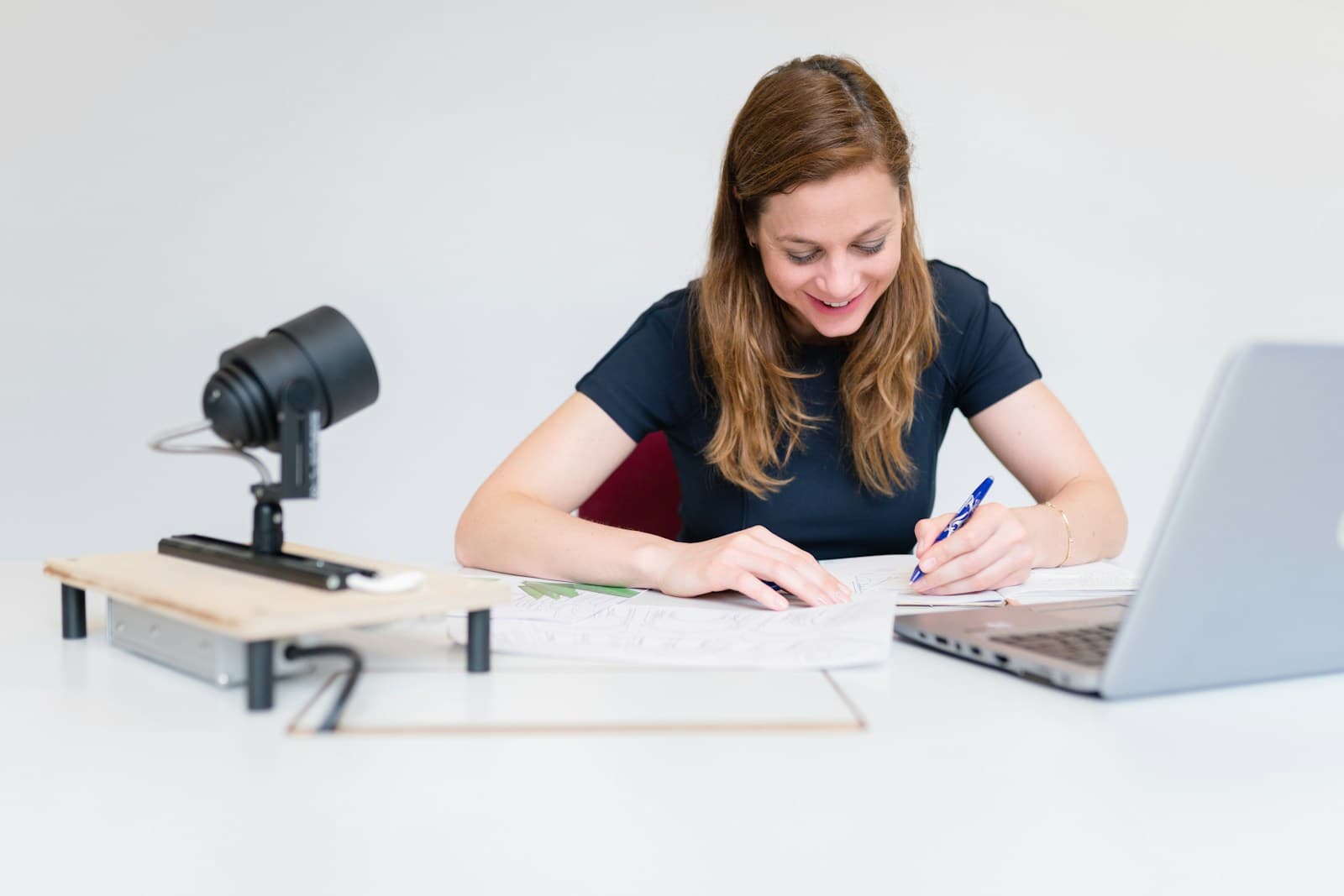 Woman writing at white desk with laptop and camera, appearing to work on documents or notes