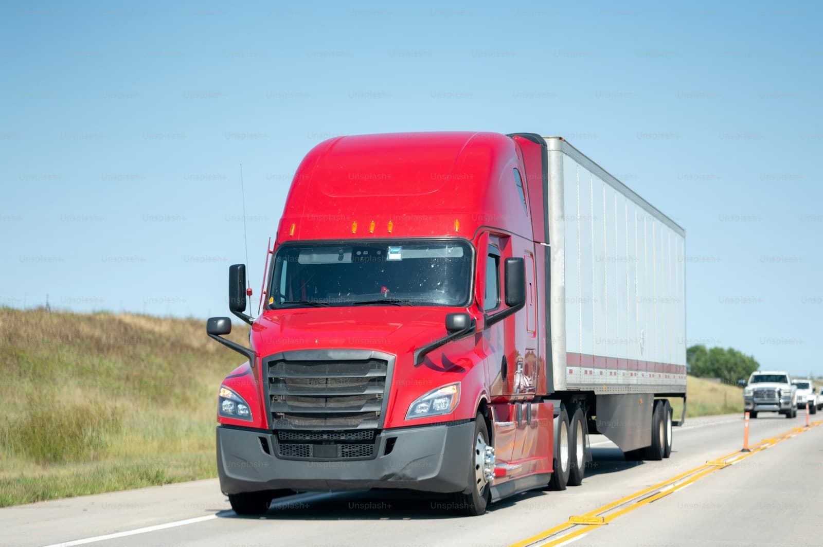 Red semi-truck with a white trailer on a rural highway under a clear blue sky