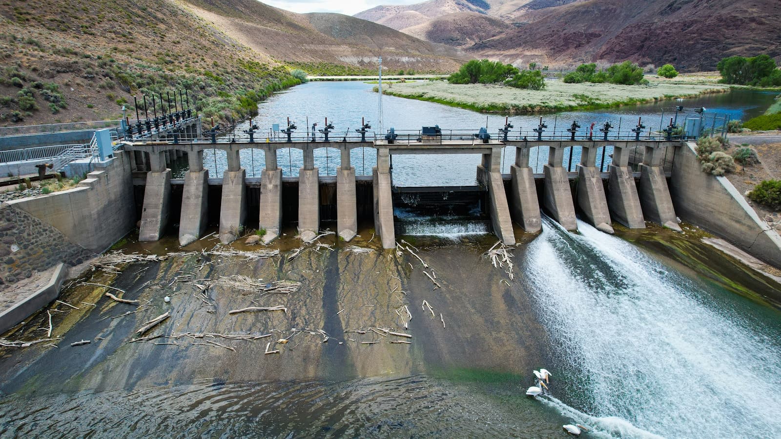 Aerial view of concrete dam with spillway gates releasing water into river in mountainous desert landscape