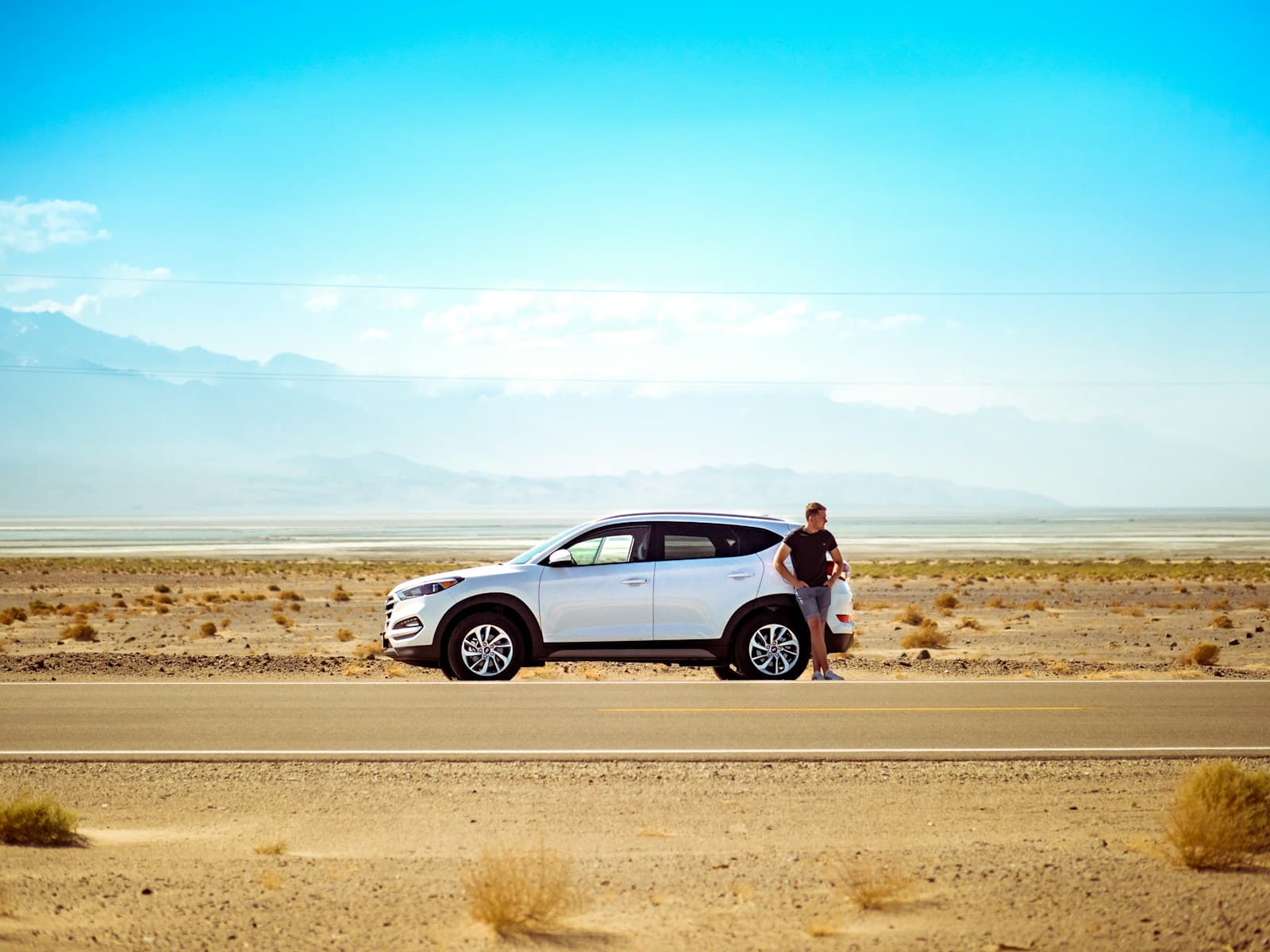 Person standing beside a white SUV on an empty desert highway below distant mountains