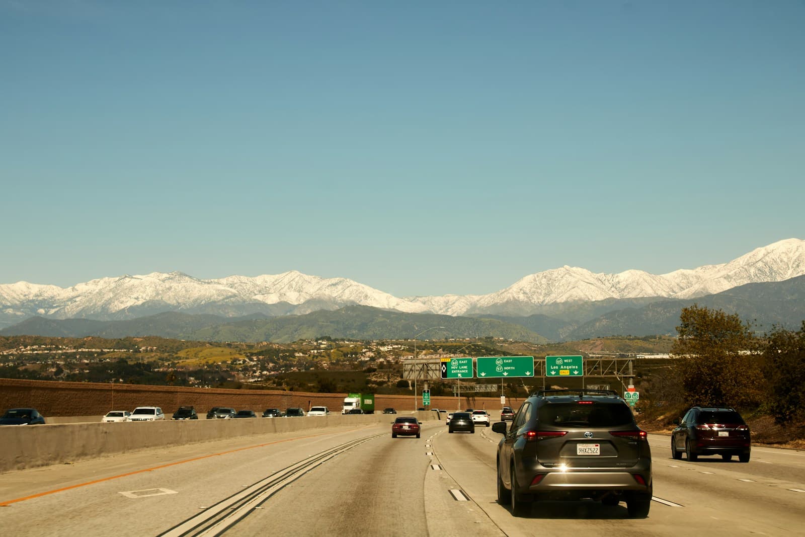 Highway traffic driving toward snow-covered mountains with green road signs overhead on a clear day
